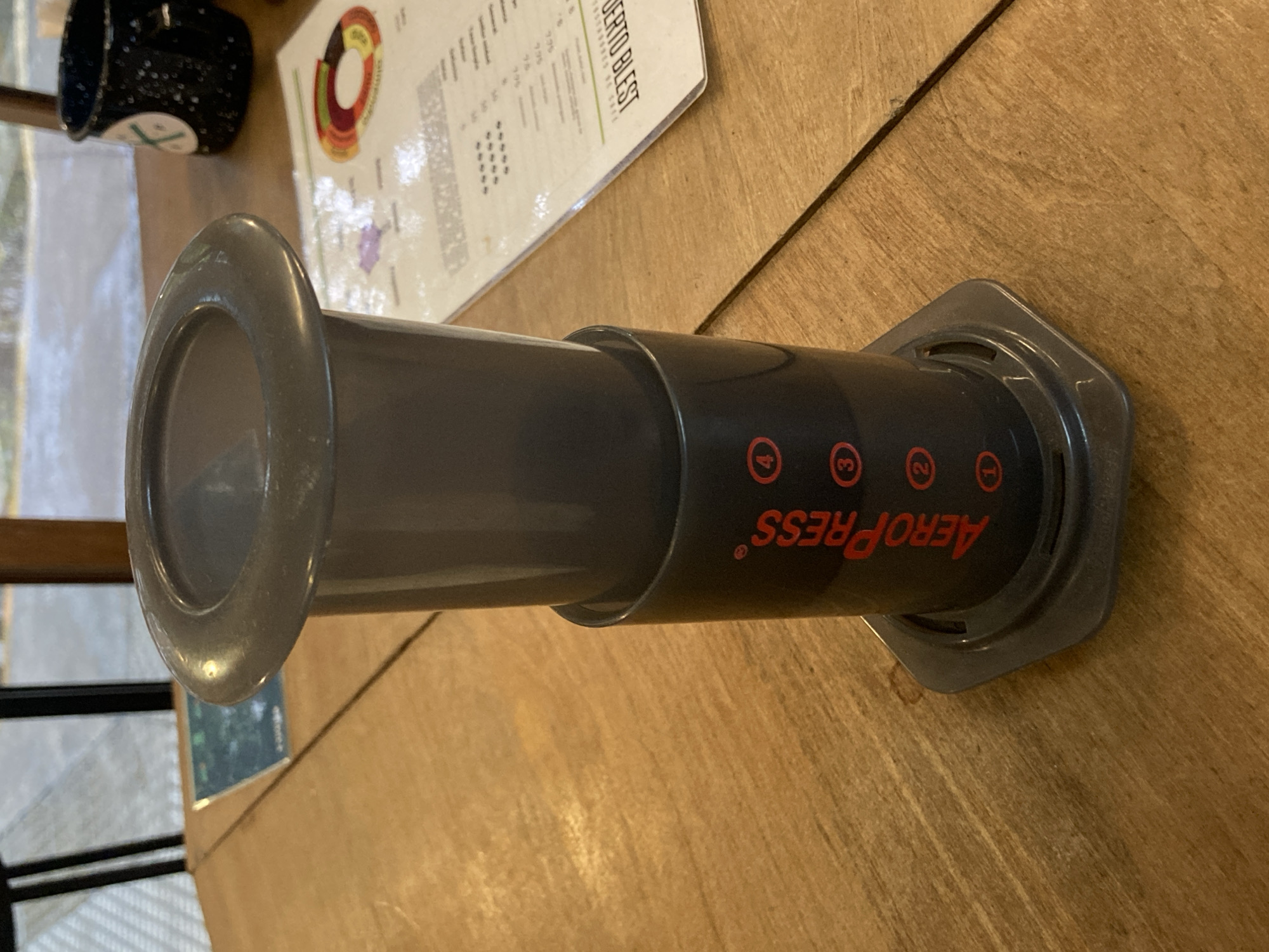 An overhead shot captures a dark gray Aeropress coffee maker resting horizontally on a light brown wooden table. The Aeropress features red branding that reads "AeroPress" and numbered circles "1," "2," "3," and "4," all appearing upside down due to the image's rotation.
In the background, a black speckled mug with a white circular logo (partially visible, showing a cross or 'X' design) sits near a white sheet of paper. This paper, possibly a menu or information sheet, contains text with partially visible words like "UERTO BLEST" and what appear to be price points such as "995." A colorful, circular diagram resembling a coffee flavor wheel is also visible on the paper.
The setting appears to be indoors, likely a cafe or a home, bathed in natural daylight, as evidenced by reflections of an outdoor scene (possibly windows and foliage) on the table surface and the Aeropress itself. No specific activity is occurring, and no people are visible. The overall impression is one of a prepared or paused coffee setup.