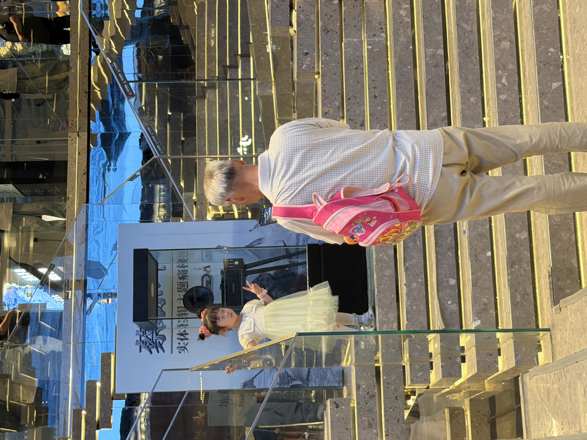In the city of Yuzhong District, China, a young girl in a light yellow dress and white tights stands on a staircase, posing with her fingers in a peace sign. She has her hair pulled up with pink bows. Beside her, an older man wearing a checkered shirt and tan pants stands with his back to the camera, holding a smartphone and looking towards the girl. He carries a pink backpack. The staircase is made of stone with integrated yellow lighting. A glass railing separates the stairs from the area where the girl is standing. The background features a reflective, mirrored wall with a display behind the girl, and more stairs ascend upwards. The overall scene appears to be an indoor public space, possibly a shopping mall or a modern building.