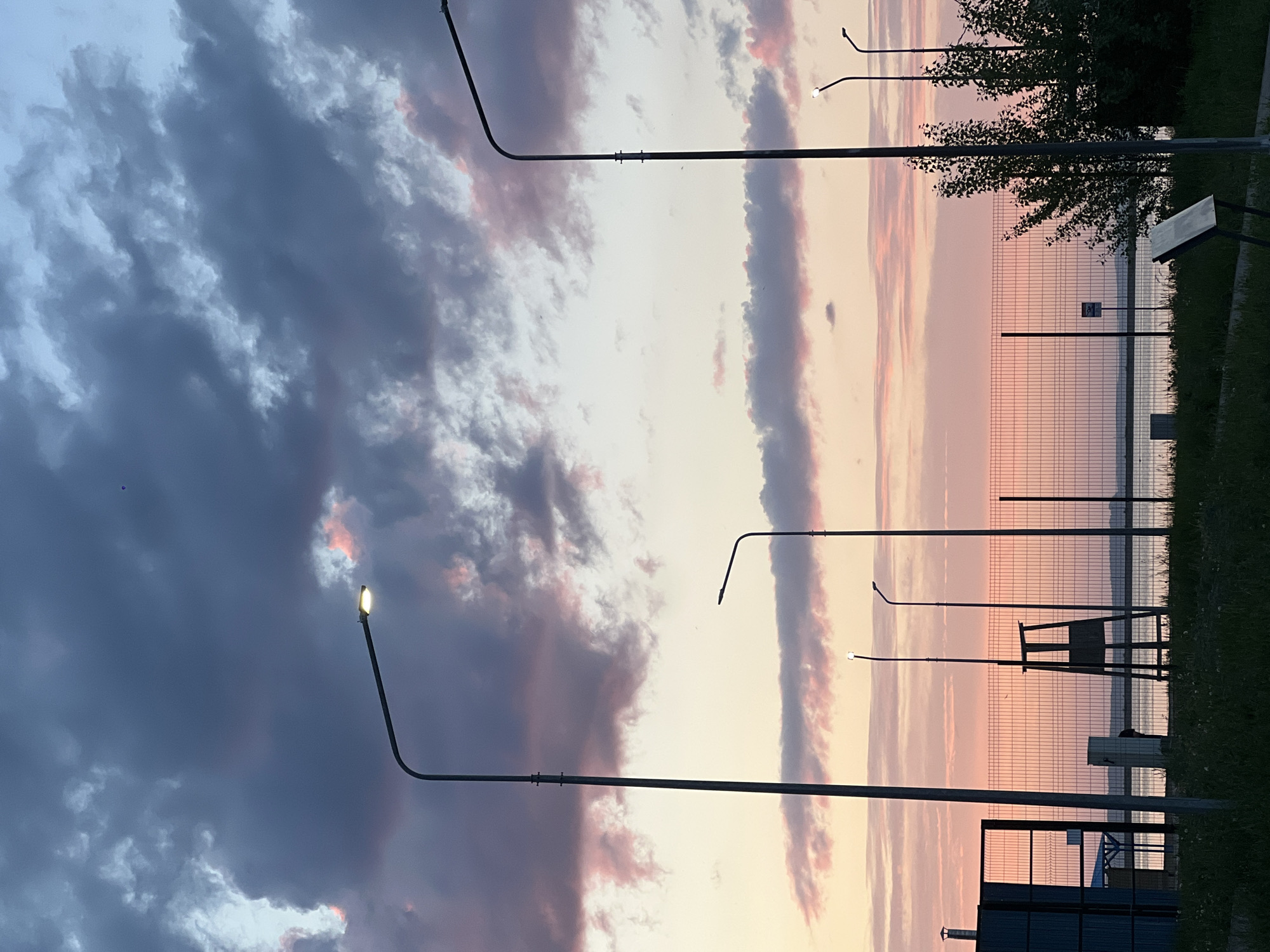 The image depicts an outdoor scene, likely near a body of water, at dusk or dawn, evidenced by the sky's gradient of pink, white, and dark gray clouds. Several tall, slender streetlights are positioned throughout the scene, with one illuminated. A wire fence runs along the horizon line, separating the foreground from the water. Behind the fence, there are indications of vegetation and possibly trees. A few structures, possibly benches or signage, are visible near the fence. The photograph appears to be taken in a public space, though no people are present. The image appears to be rotated 90 degrees counter-clockwise. The location is specified as Chaikovsky, Russia, though nothing directly in the image suggests this location.