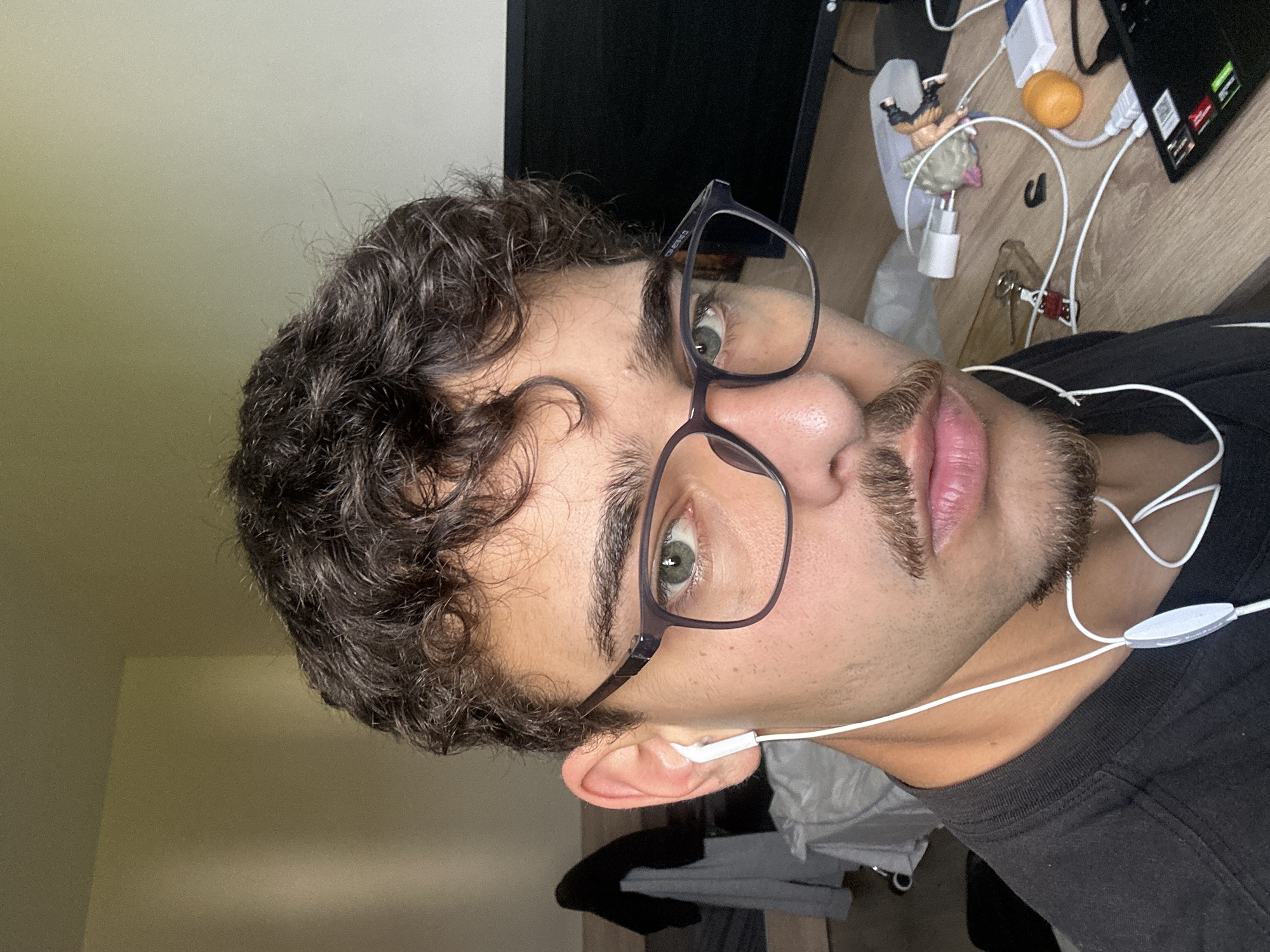 A portrait of a young adult male wearing glasses and earphones is presented, rotated 90 degrees counter-clockwise. The man has curly dark hair and a patchy beard. He is wearing a black t-shirt. In the background, there is a wooden desk with various objects including a computer monitor, keyboard, cables, a small figurine, and an orange. The environment appears to be indoors, likely a home office. The city of Sofia, Bulgaria is not discernible from the image.