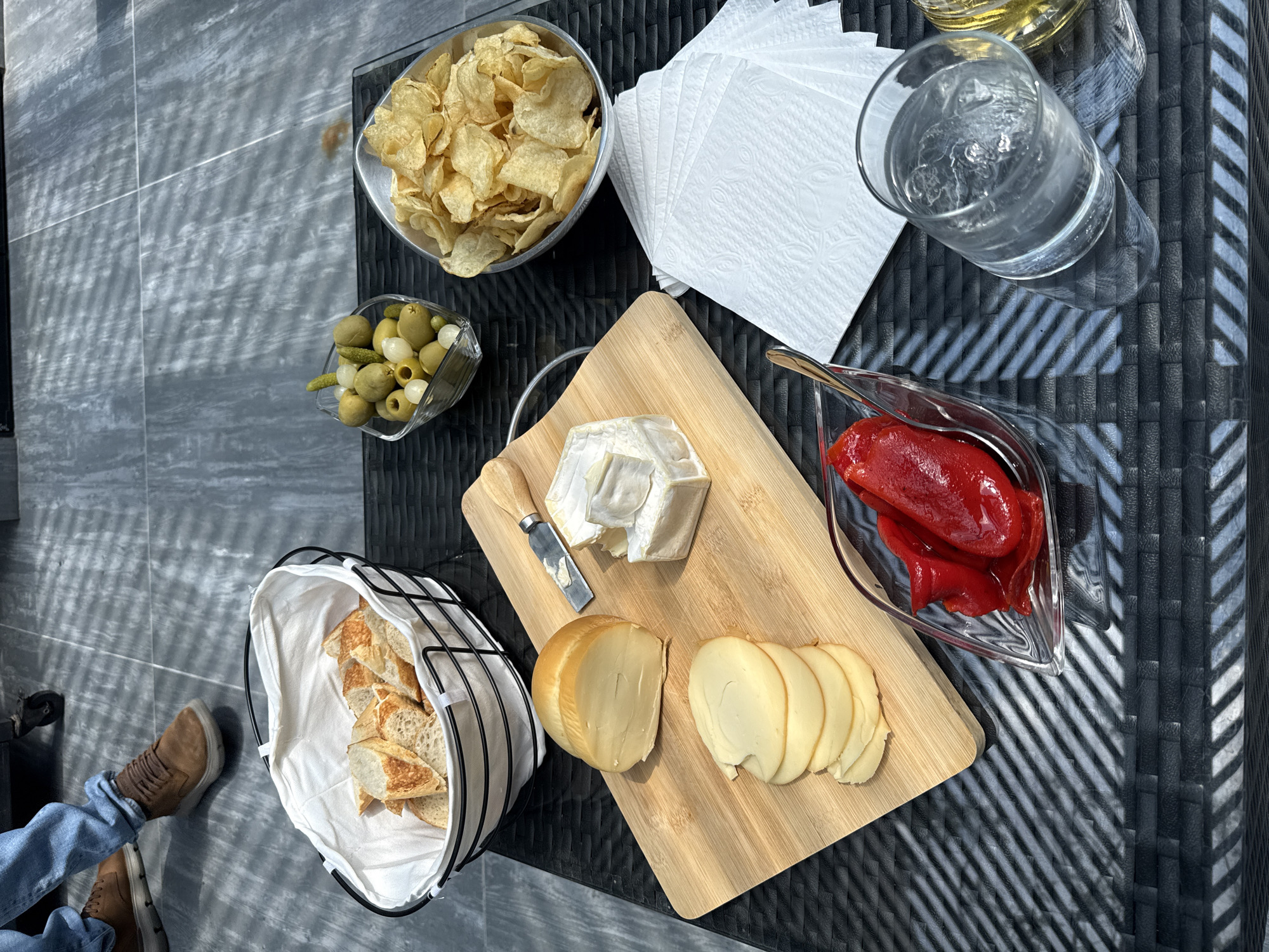 An overhead shot captures a casual outdoor snack spread on a dark, textured table, likely a patio or garden setting in Huixquilucan de Degollado, Mexico. The scene is brightly lit by direct sunlight, casting sharp, distinct shadows, suggesting it is a clear, sunny day, likely late morning or afternoon.
Various food items are arranged on the table:
- A large metal bowl filled with potato chips is in the upper left.
- Below it, a glass bowl holds green olives, small white pickled onions, and cornichons.
- A wooden cutting board, centrally placed, features a wedge of Brie or similar soft cheese with a small knife, and a round, light brown smoked cheese, partially sliced.
- To the right of the cutting board, a glass dish contains vibrant red roasted peppers.
- In the lower left, a basket lined with white cloth holds several slices of baguette or similar bread.
- A stack of white embossed paper napkins is placed near the chips.
- Two glasses are visible in the upper right: one filled with ice water, and another, partially visible behind it, possibly containing a light-colored beverage.
In the bottom left corner, the lower legs and feet of a person are visible, clad in blue jeans and brown shoes, resting on the grey tiled floor, suggesting a relaxed posture. No visible text is present in the image. The arrangement implies a leisurely moment enjoying food and drink outdoors.
