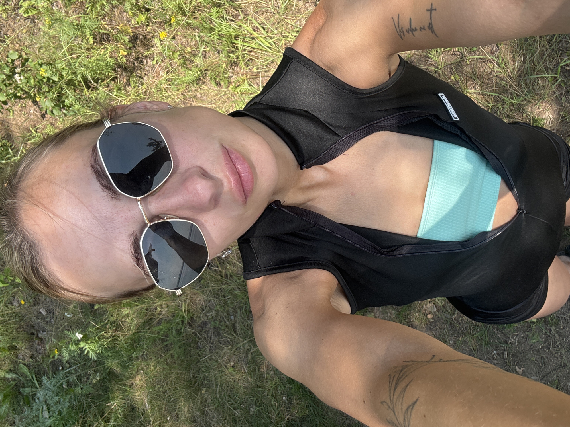 A woman with fair skin and brown hair, wearing sunglasses, a black sleeveless top, and a light blue bra, poses for a selfie outdoors. She has tattoos on her arms and is standing in a grassy area with some yellow flowers. The scene is set in the city of Noviy, Russia.