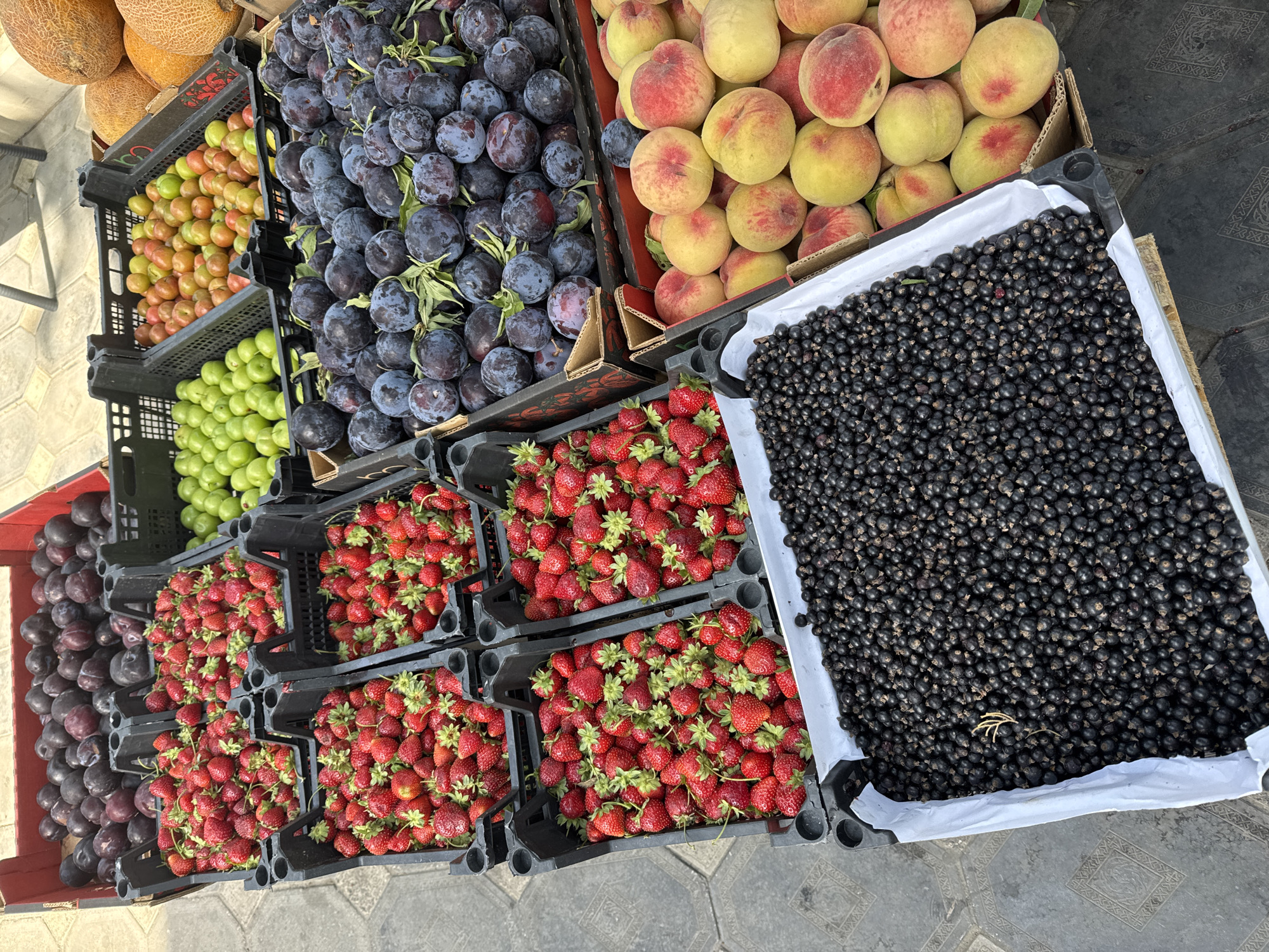 This image shows an outdoor fruit stand in Baku, Azerbaijan, with an assortment of fresh produce displayed in plastic crates and cardboard boxes. The fruits visible include plums, strawberries, green plums, and peaches. In the foreground, a large crate is filled with black currants. The ground beneath the fruit stand is paved with textured tiles. The scene appears to be a market setting, with various fruits laid out for sale. There are no people or animals visible in the image. The lighting suggests it is daytime.