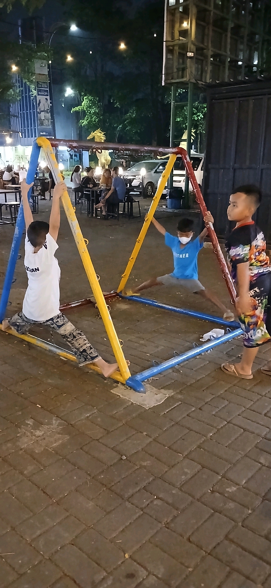 Three young boys are playing on a colorful playground structure, possibly a monkey bar or climbing frame, in an outdoor setting at night. The structure is made of red, yellow, and blue metal bars. The boy on the left, wearing a white t-shirt and patterned pants, is standing with his arms raised, holding onto the blue and yellow bars. In the center, a boy in a blue t-shirt and gray shorts is in a wide stance, with his hands on the red and yellow bars. He is wearing a white face mask. To the right, a boy in a black t-shirt with a colorful graphic print and patterned shorts stands and observes.
The ground is paved with interlocking dark-colored bricks, arranged in a herringbone pattern. In the background, there are trees and streetlights, suggesting an urban environment. Behind the playground, there are tables and chairs, indicating a seating area, possibly for a food establishment. A white vehicle is visible in the background, and a tall, multi-story structure with wooden beams and lights is also present. The lighting appears to be artificial, creating a warm glow in some areas and casting shadows.
The location is identified as Taman Ganesa, which is part of the city of Cirebon, Indonesia. Taman Ganesa is a public park located in front of the Cirebon City Hall, known for its community activities and green spaces.