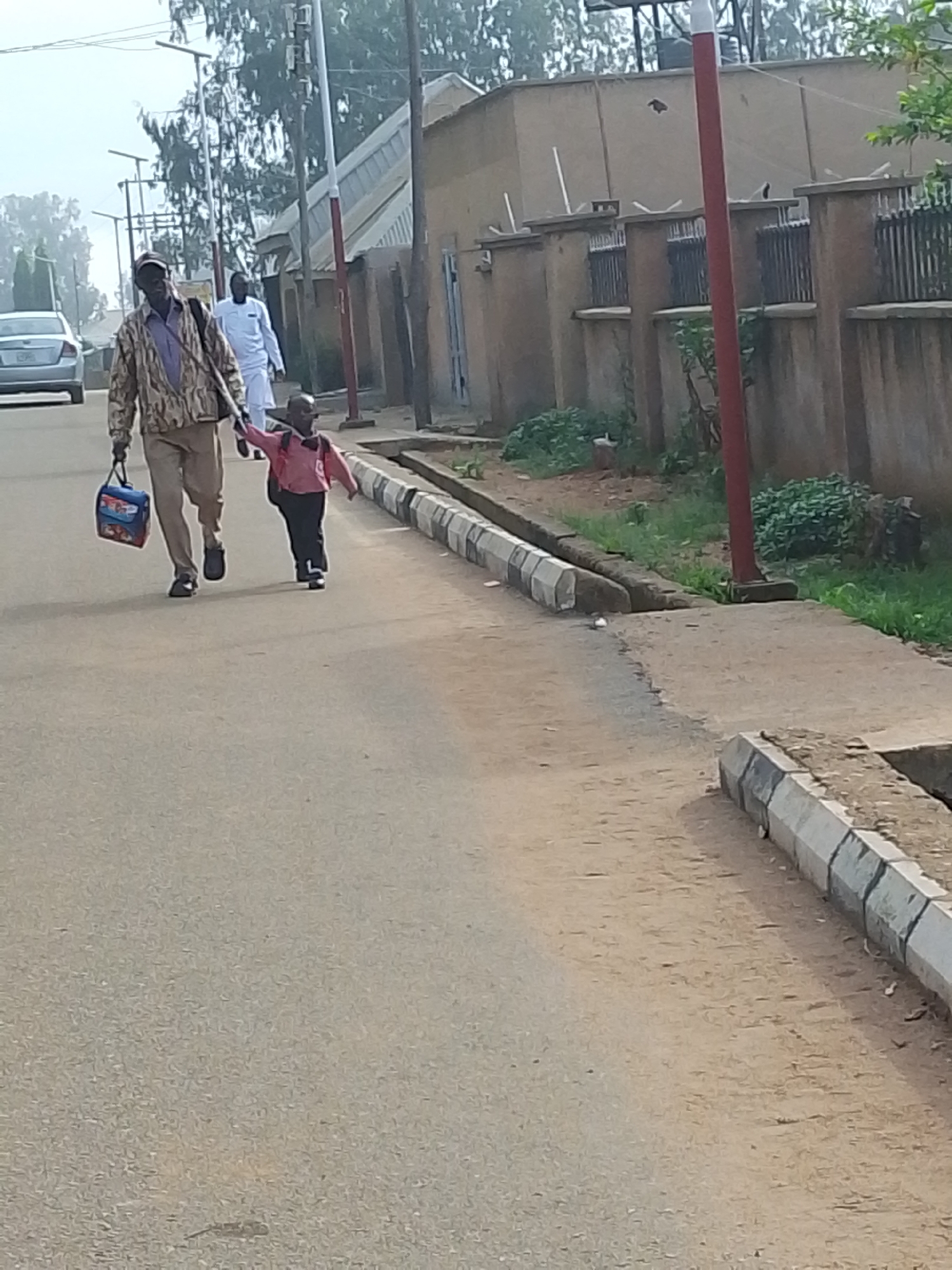 The image depicts an adult male and a child walking hand-in-hand on a street in Jos, Nigeria. The adult male, dressed in a patterned jacket and trousers, is carrying a blue lunchbox in his left hand. The child, dressed in a pink shirt and dark trousers, is holding the man's right hand. A second adult male, dressed in a white robe, is walking behind them.
The setting is a residential street lined with buildings and a concrete wall topped with barbed wire. A car is parked on the side of the street. A curb runs along the edge of the road, separating it from a dirt or grassy area. A red pole stands on the grassy area adjacent to the curb.
The overall environment suggests a sunny day in a residential area within Jos, Nigeria.