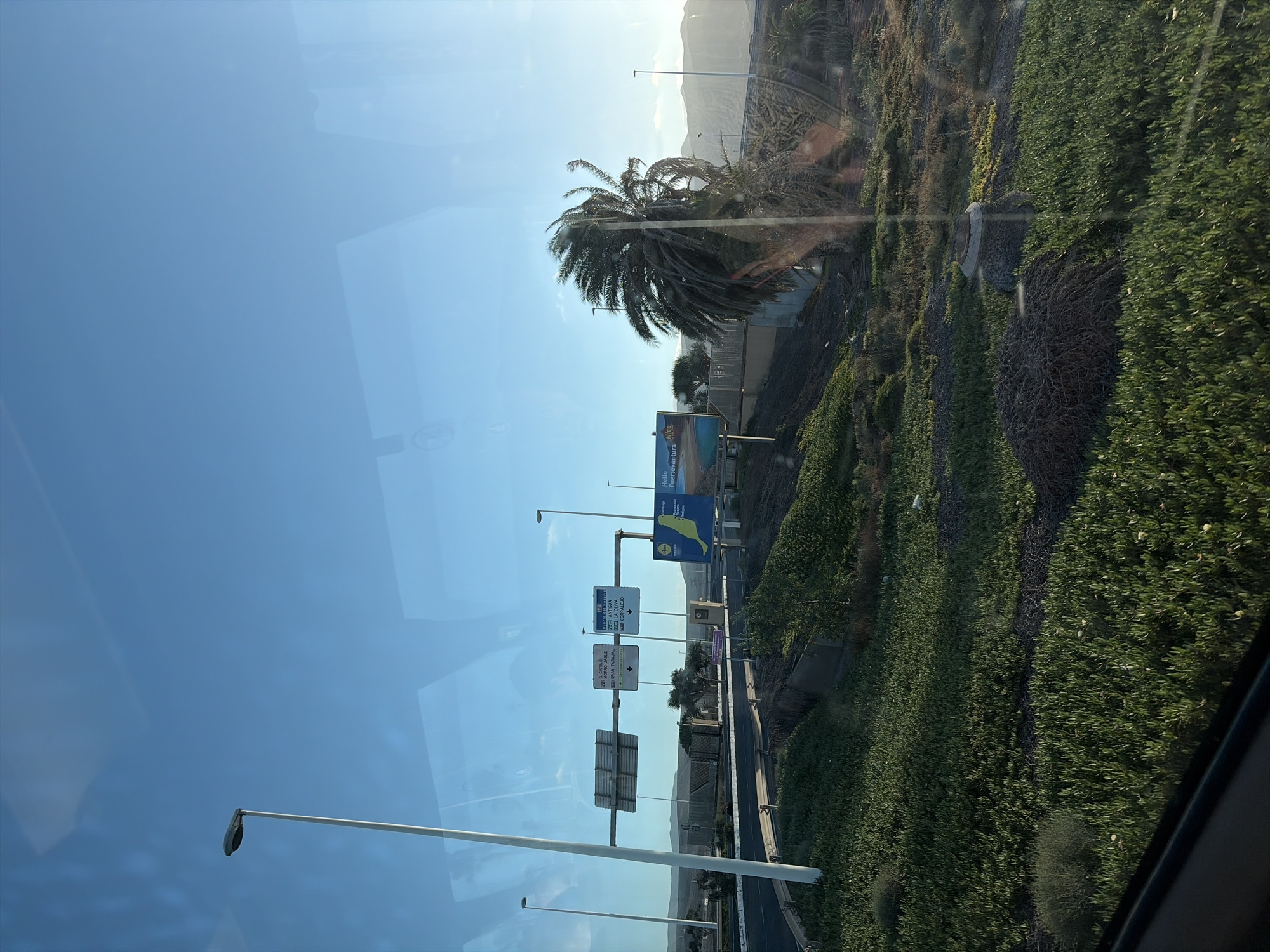 The image shows a landscape in Puerto del Rosario, Spain, observed through a window. A palm tree stands next to the road. Street signs indicate directions to Morro Jable, Gran Tarajal, Puerto del Rosario, Antigua, La Oliva, and Corralejo. A billboard advertises Fuerteventura. The environment features a mix of greenery and mountainous terrain in the background. The sky is clear and blue.