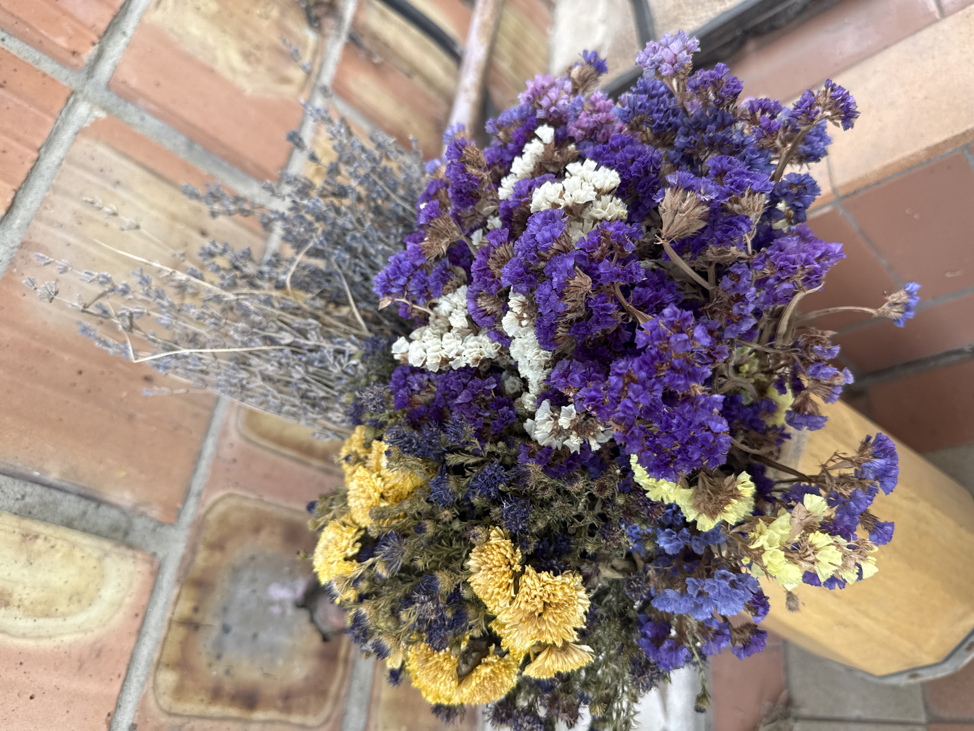 A close-up, overhead view captures a bouquet of dried flowers in various colors, including yellow, purple, white, and lavender. The flowers are arranged in a wooden container, partially visible at the bottom of the frame. In the background, a textured surface of red-orange bricks with gray grout is visible, suggesting an outdoor or rustic setting. The lighting is natural, casting subtle shadows and highlighting the delicate textures of the dried flora. There are no people or animals present in the image. The scene is static and focuses on the arrangement of the flowers against the brick backdrop. The location is identified as Obukhivka, Ukraine, implying this is a snapshot of a scene within that city.