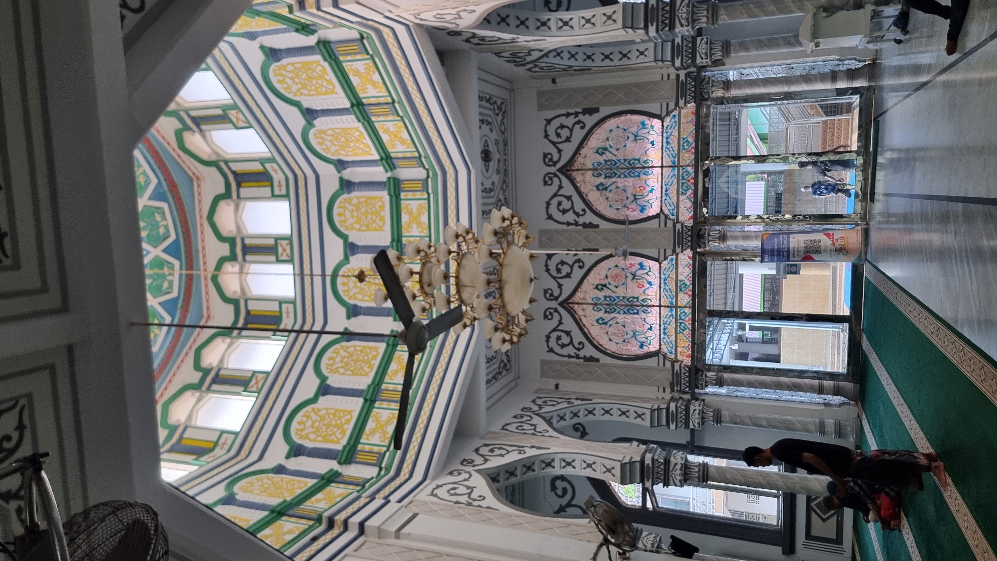 An interior view of a mosque in Keudekruenggeukueh, Indonesia, captures the grandeur of its architecture and the serene atmosphere. The camera is positioned low, looking upwards towards a domed ceiling adorned with intricate colorful patterns in blue, green, and yellow. A large, multi-tiered chandelier hangs from the center of the dome, casting a warm light.
The walls of the mosque are decorated with white and grey motifs, featuring arched doorways and stained-glass windows in pink and blue hues. A large ceiling fan is visible, suggesting the ambient temperature.
In the foreground, a green carpet with decorative borders covers the floor, indicating a prayer area. Several individuals are present. One man, wearing a black t-shirt and a sarong, stands facing away from the camera. Two other figures, partially visible, are seated on the carpet.
The far end of the mosque opens into a brighter area with glass doors, revealing a staircase and more architectural elements. A banner with a QR code and text in Indonesian is placed near the entrance. Another man is seen walking through the glass doors, and a third figure is visible further in the distance. The flooring in the open area appears to be polished stone or tile. The overall scene conveys a sense of religious sanctity and architectural beauty.