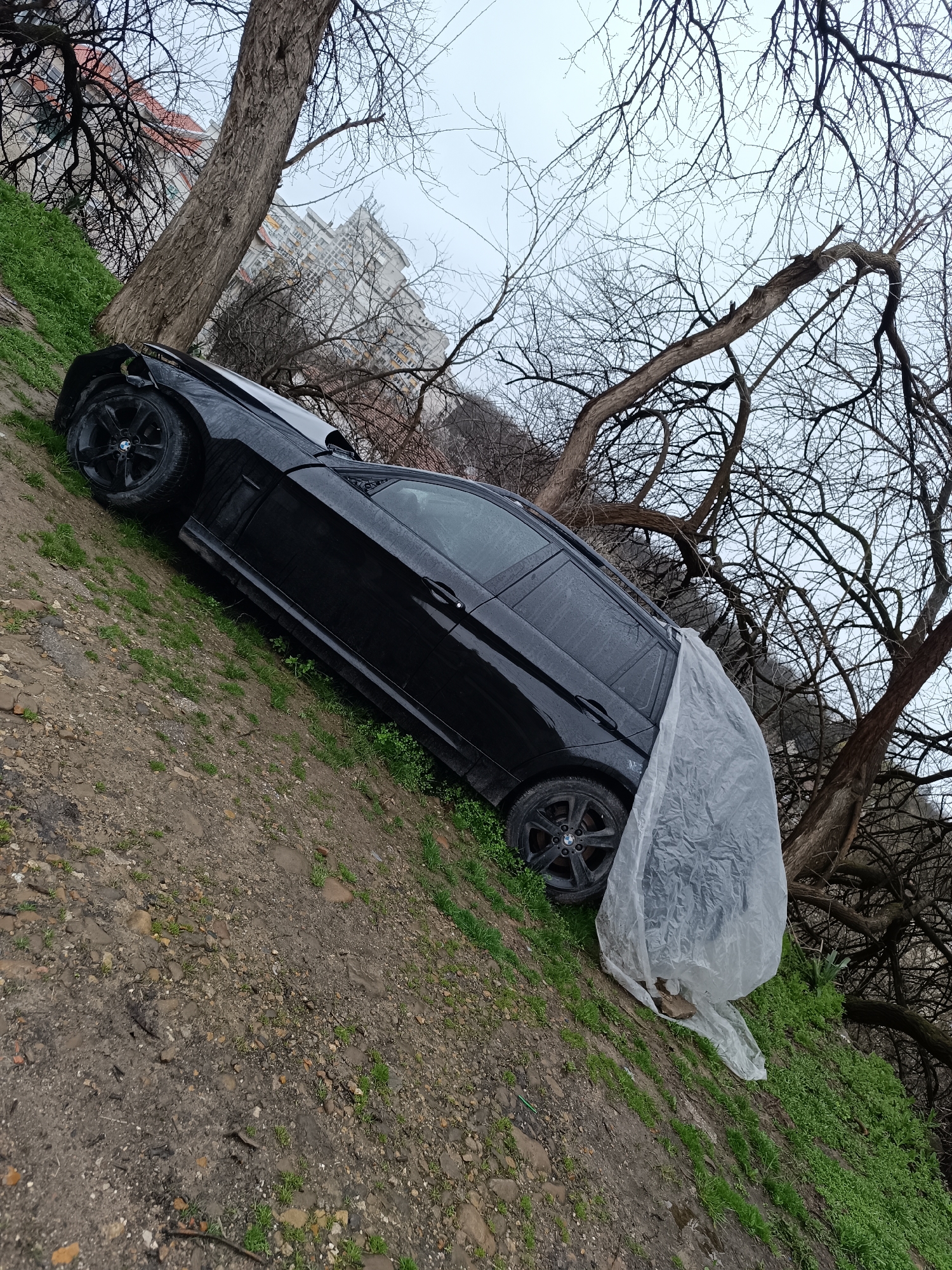 A black BMW SUV is situated on a grassy and muddy slope beneath trees. The front left of the vehicle is damaged, and a translucent plastic sheet covers the rear driver's side. The ground is damp with scattered stones and sparse vegetation. Bare tree branches frame the scene against a cloudy sky. In the background, there is a multi-story building, indicative of an urban environment. The location is likely in Pleven, Bulgaria.