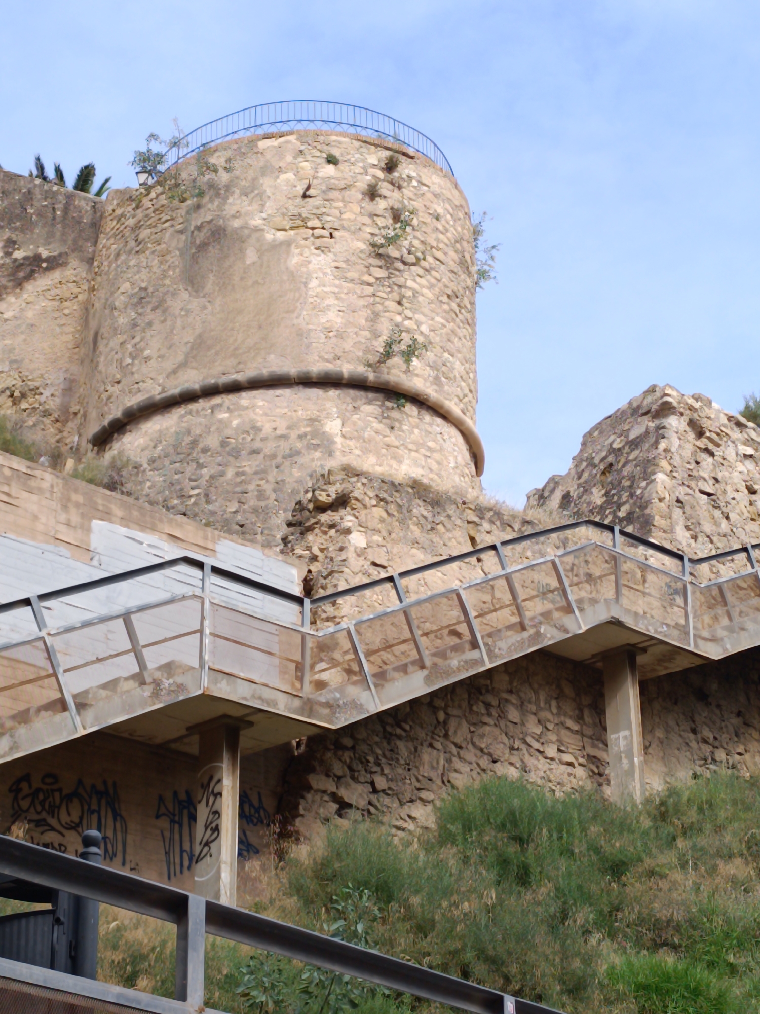 A low-angle, outdoor shot captures a section of an ancient stone structure, possibly a castle or fortress, against a bright blue sky. The stone is weathered and rough, with patches of vegetation growing on its surface. To the right of the stone structure, a modern staircase with metal railings and glass panels ascends. The staircase is constructed over a concrete wall that has graffiti in blue spray paint. The surrounding environment is overgrown with green bushes and dry grass. The scene appears to be a public space, possibly a historical landmark with public access. The time of day is daytime, indicated by the bright sky and clear lighting.
