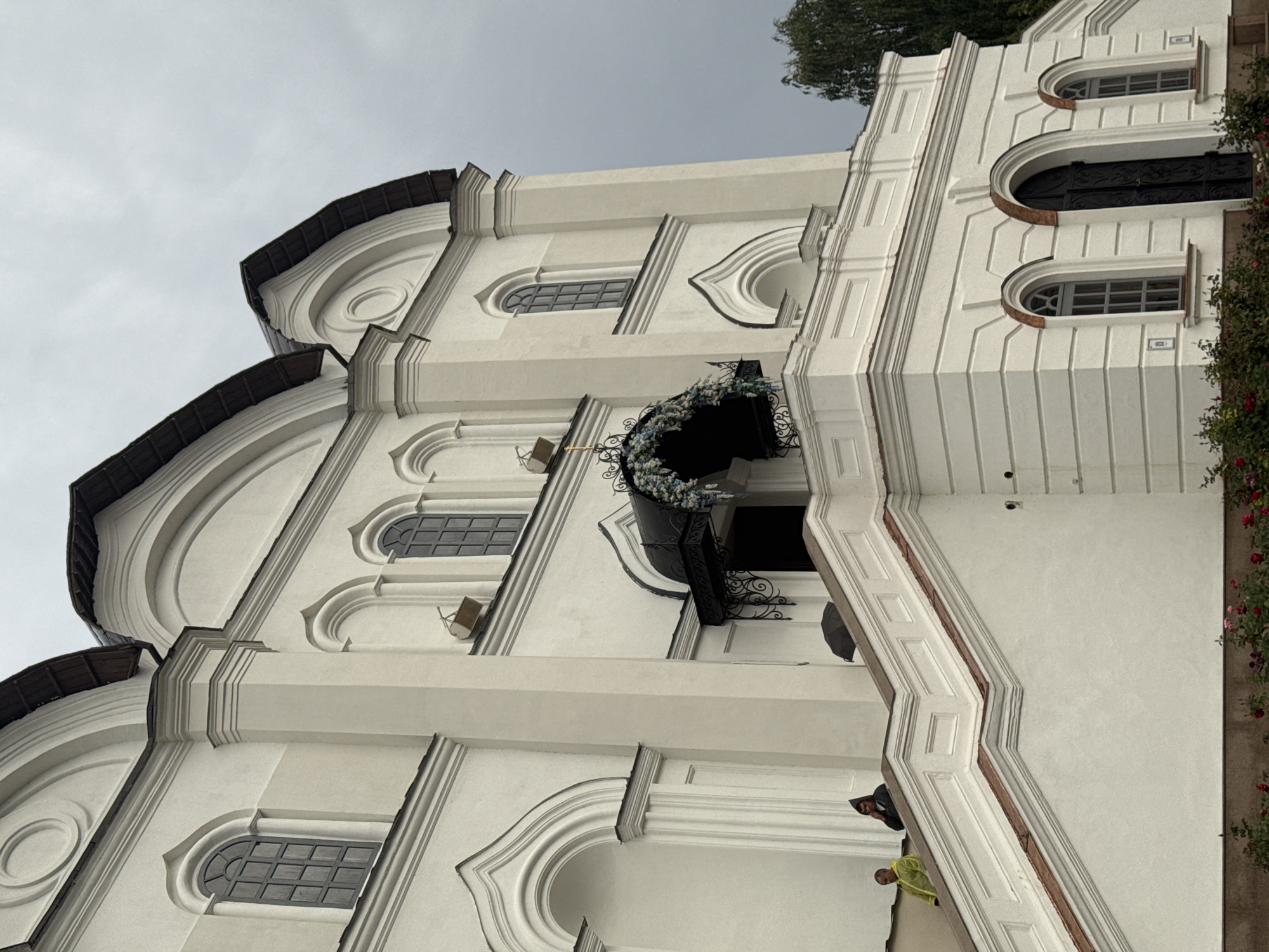The image shows a portion of the exterior of the Annunciation Cathedral in Kazan, Russia, under a cloudy sky. The cathedral is a large, white, multi-domed structure with arched windows and ornate architectural details. In the foreground, a wide white staircase with red trim leads up to a balcony. Two men are visible on the staircase: one on the left is wearing a bright yellow jacket, and another to his right is wearing a dark hooded jacket and holding a cell phone to his ear. An open black umbrella rests on the railing of the staircase. In the distance, a lush green tree is visible to the right of the cathedral. The base of the cathedral is surrounded by blooming rose bushes.