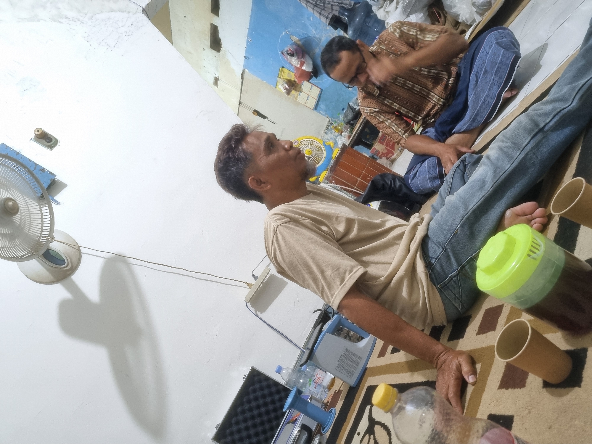 In Prambon, Indonesia, two men are seated on a patterned carpet in what appears to be a dimly lit room. The man in the foreground, wearing a light brown t-shirt and blue jeans, is leaning back on his left hand, looking towards the right. To his right, a second man, dressed in a brown and white patterned shirt and blue sarong, is sitting cross-legged, with his right hand touching his face. He is looking downwards.
Several objects are scattered around the men. To the left of the foreground man is a partially visible clear plastic bottle with a yellow cap and a cylindrical brown paper cup. Next to these is a green and clear plastic container with a green lid. Further to the left, on a table or raised surface, are two bottles of water, a blue object, and a black case with a textured interior. Behind the seated men, there is a blue wall with various items attached and leaning against it, including a yellow fan and a water cooler. A white wall with a ceiling fan is visible in the upper left portion of the image, casting a large shadow on the wall. Electrical cords are running from the ceiling fan and other devices. The overall impression is of a casual gathering in a domestic or informal setting.