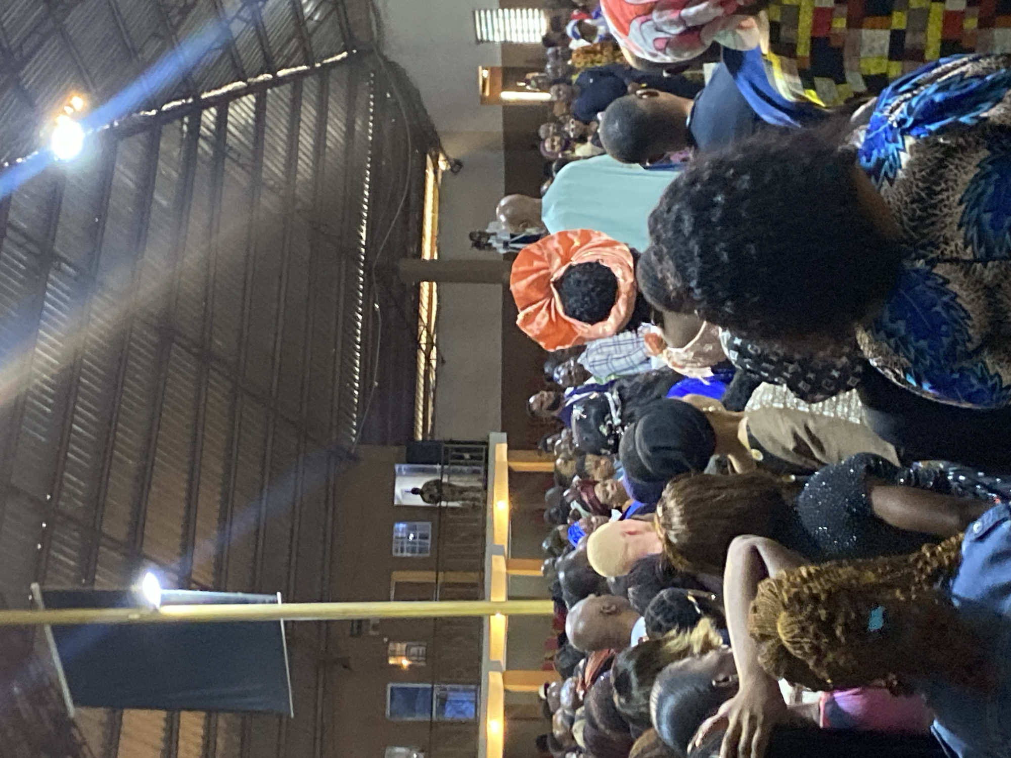 The image depicts a crowded indoor gathering, likely a church service or community event, located in Rukuba Road, Nigeria. A large group of people, predominantly Black Africans, are seen from behind, facing towards a stage area or a focal point at the front of the room. The architecture suggests a large hall with a corrugated metal roof and exposed beams, illuminated by overhead lighting and a prominent beam of light streaking from the upper right. In the background, there is a balcony with people standing and looking down at the gathering. The individuals in the foreground are adorned in vibrant clothing and headwraps, common in Nigerian cultural attire. The overall atmosphere appears to be one of collective attendance and engagement.