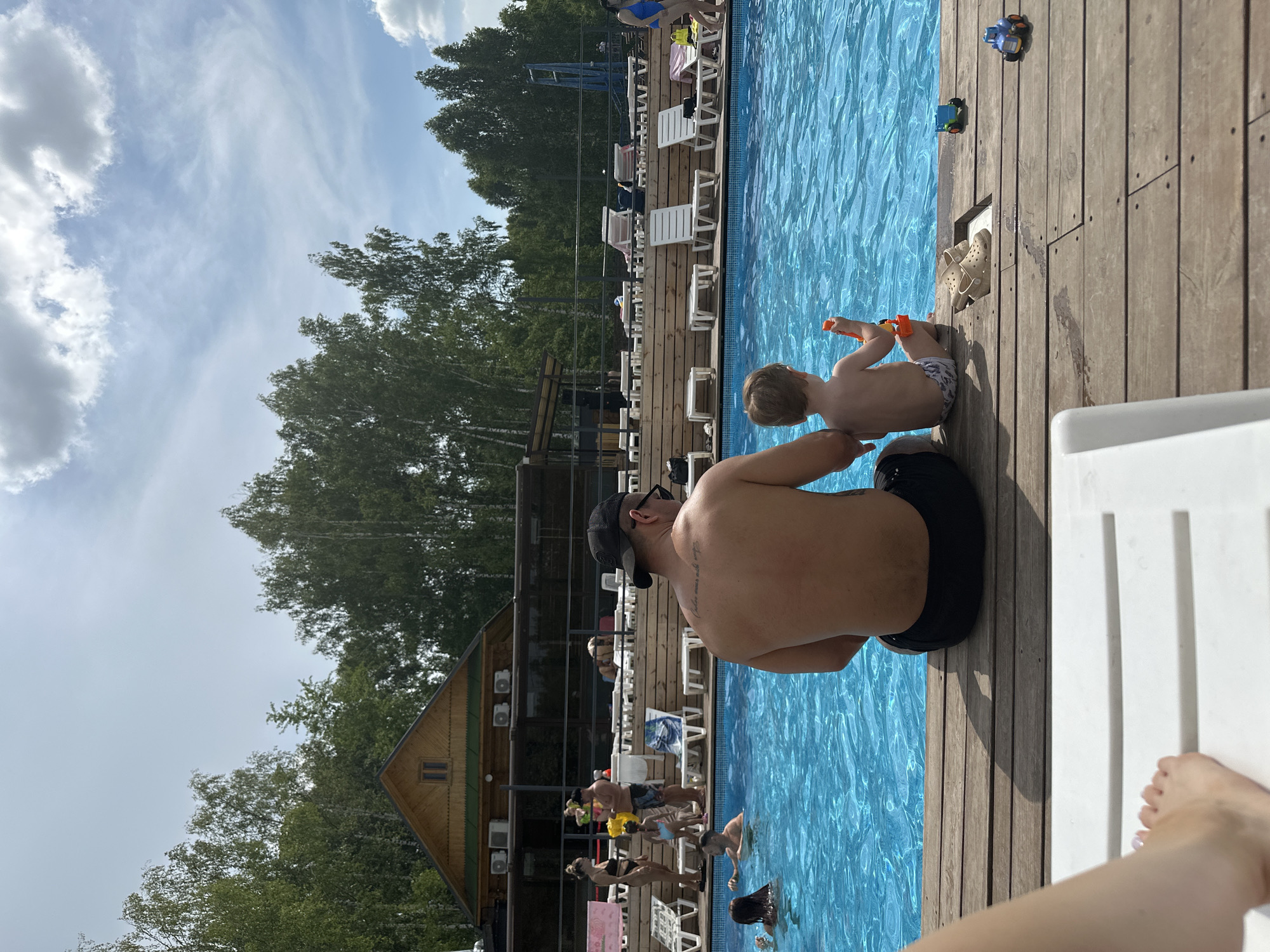 This is a photograph of a swimming pool area, likely a resort or water park, with a wooden deck in the foreground. In the center of the image, a shirtless man wearing black swim trunks and a black baseball cap is seated with his back to the camera, facing a young child. The child, also shirtless, is seated next to him, facing away from the camera, and holding an orange toy. The pool is filled with bright blue water, and several people are visible swimming and relaxing in and around it. In the background, there is a wooden building with a pitched roof, surrounded by trees. The sky is partly cloudy. There are no visible landmarks that specifically place this location within the city of Kazan, Russia.