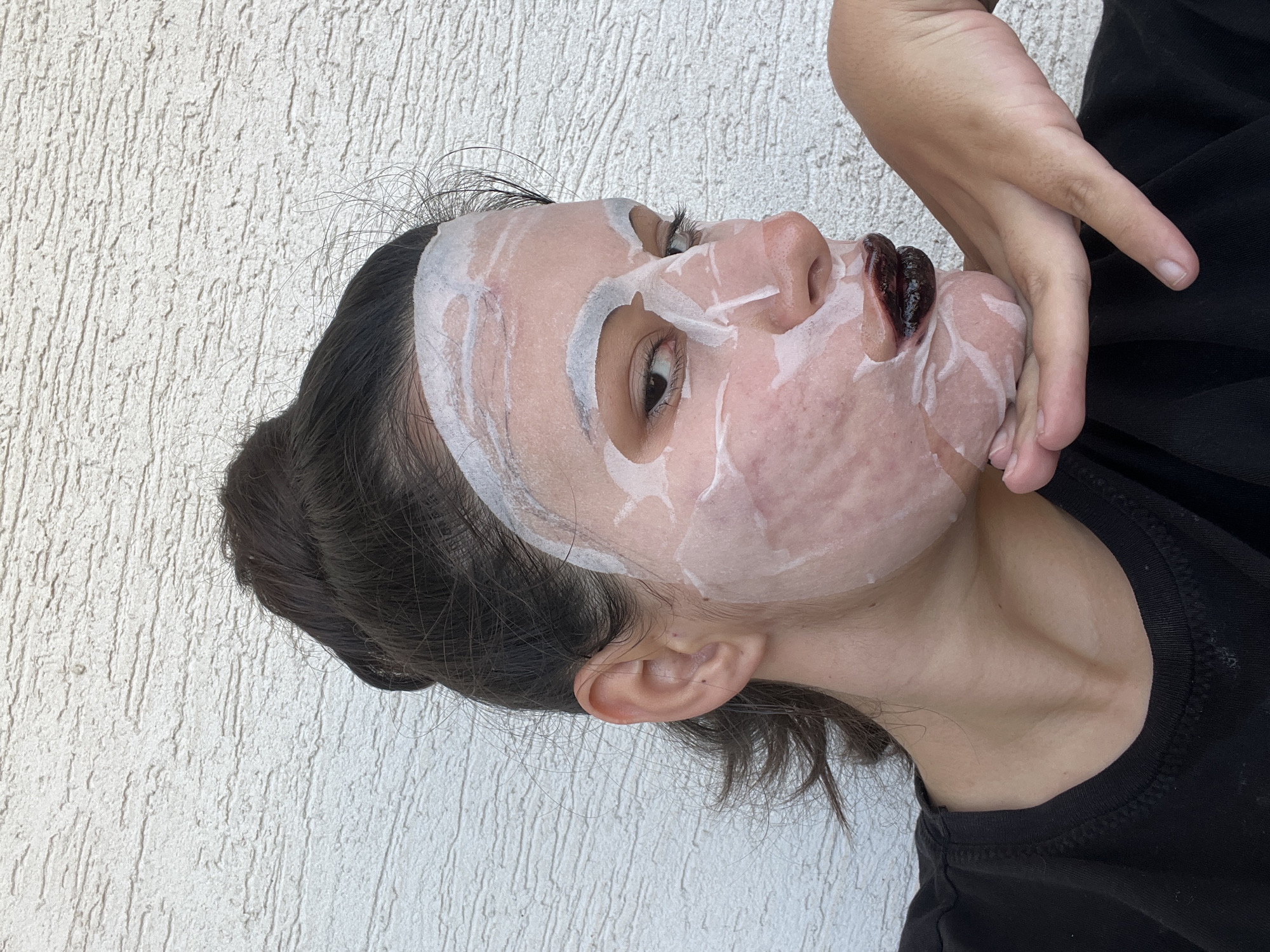 A person wearing a white face mask and dark lipstick lies on their side. The person's skin is visible beneath the mask. The person is wearing a black shirt and holding their hand near their face. The background is a white textured wall. The image does not contain sufficient information to determine that this image was taken in Kiryat Bialik, Israel.