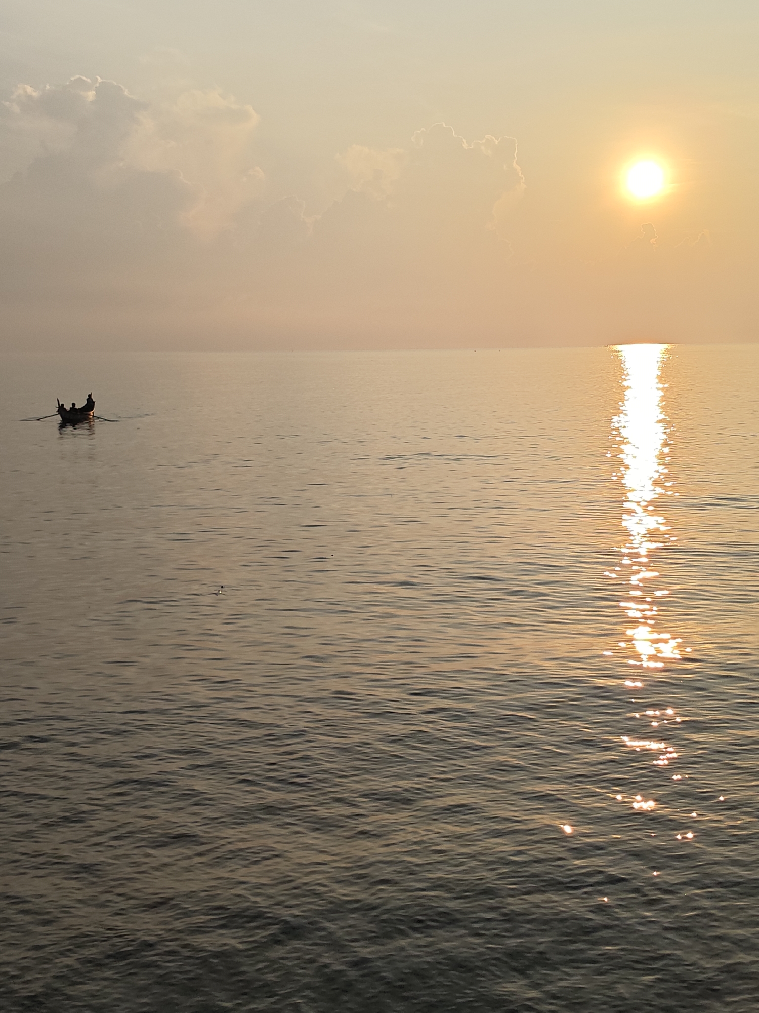 The image depicts a serene coastal scene, likely taken in Lhokseumawe, Indonesia. A small boat with two or three individuals aboard is visible in the distance to the left. They appear to be rowing or navigating through the calm water. The sun is positioned high in the sky towards the right, casting a shimmering reflection on the surface of the water that creates a striking vertical line of light. Soft clouds are scattered across the sky, some appearing near the horizon. The overall color palette is dominated by warm tones, primarily oranges, yellows, and browns, which suggests that the image was taken during sunrise or sunset. The water exhibits subtle ripples, contributing to the tranquility of the scene.