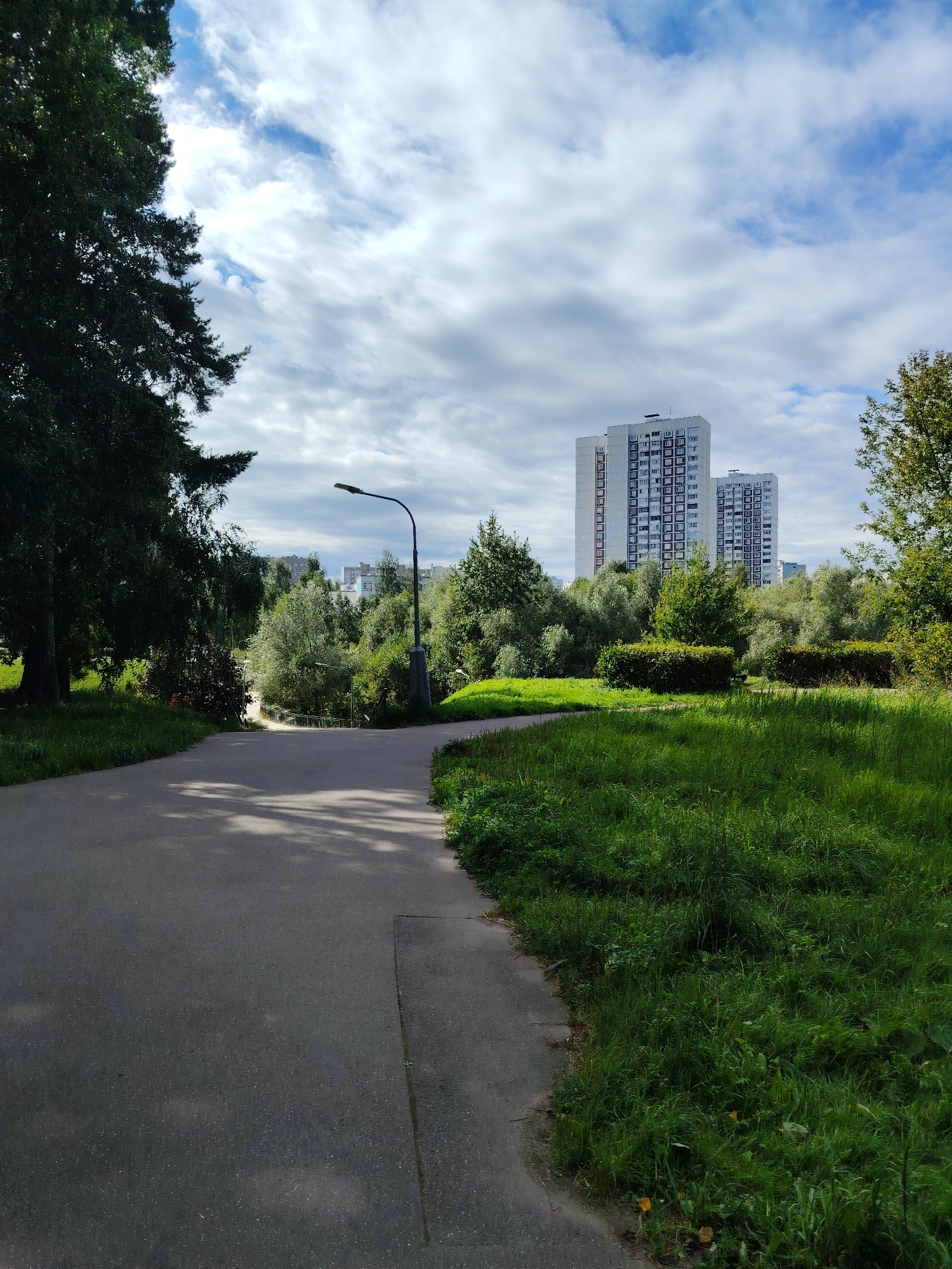 The image depicts a park pathway in Zelenograd, Russia, under a cloudy sky. A paved path curves to the right, bordered by lush green grass on the right and trees on the left. In the background, several tall, white apartment buildings with dark-framed windows are visible above a dense line of trees. A lamppost stands along the path. The scene is a tranquil outdoor setting, likely a residential area.