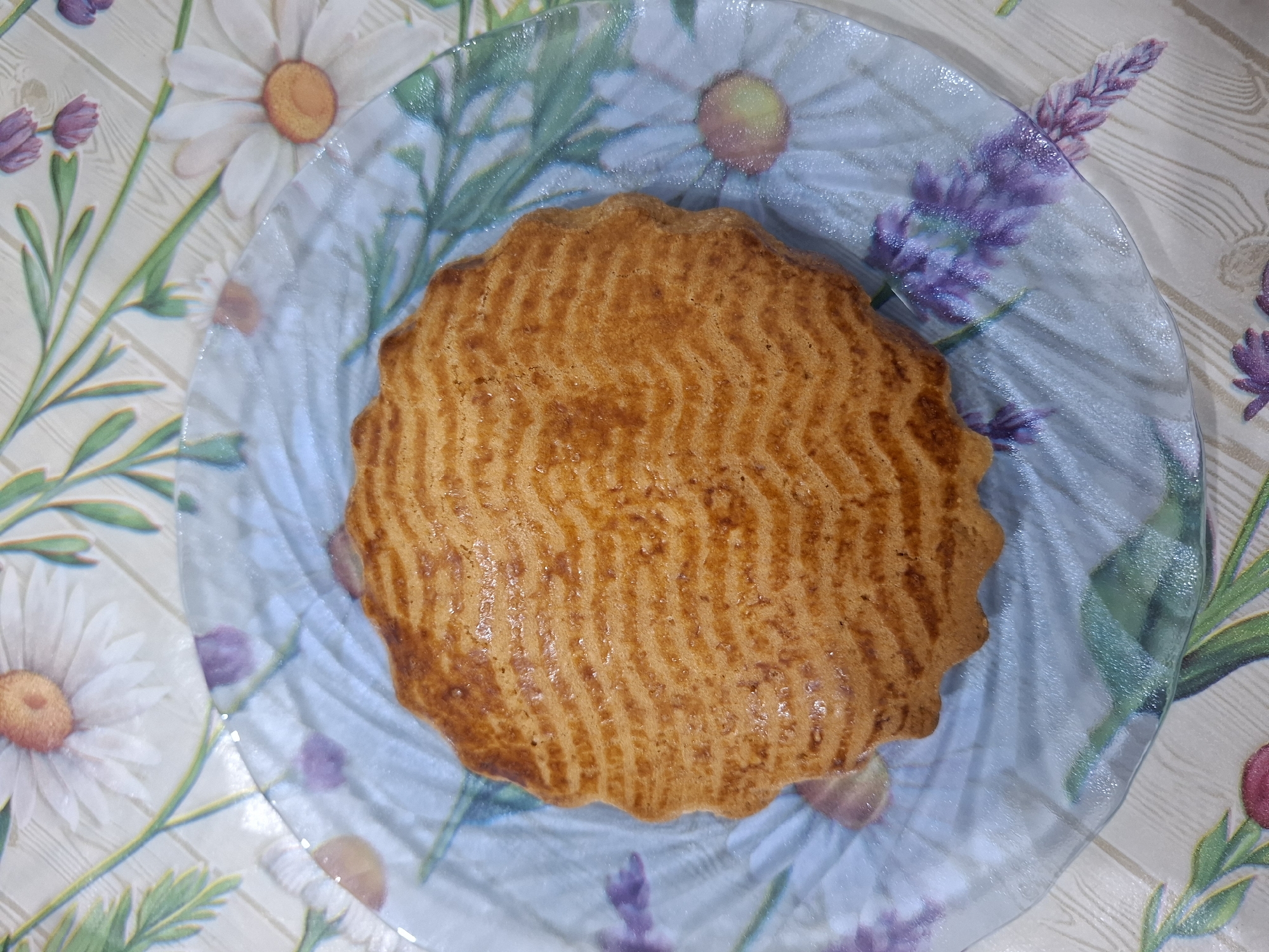 A circular, golden-brown cake with a wavy patterned crust sits on a clear glass plate. The plate is decorated with a floral pattern of white daisies and purple lavender sprigs against a light, wood-grained background. The cake itself has a slightly ruffled edge. The image is taken from a top-down perspective. The setting appears to be indoors, possibly on a table, given the tablecloth-like background pattern. No people are visible, and no text is present in the image. The lighting suggests it could be daytime.