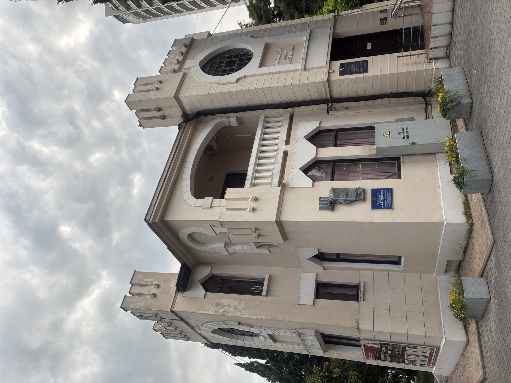 The photograph shows a light-colored, two-story building with castle-like crenellations at the roofline. The building has distinctive architectural elements such as arched windows and decorative stonework. There is a balcony with a white railing on the second floor. Several plaques and signs are affixed to the exterior walls, including one that reads "БИБЛИОТЕКА ИМЕНИ А.С. ПУШКИНА" (Library named after A.S. Pushkin). A metal utility box is situated near the entrance. Two concrete planters filled with yellow flowers and greenery are placed in front of the building. The ground in front of the building is paved with interlocking stones. In the background, a modern high-rise building and lush green trees are visible. The sky is overcast with clouds. This is the A.S. Pushkin City Library located at 13 Kurortny Prospekt in Sochi, Russia.