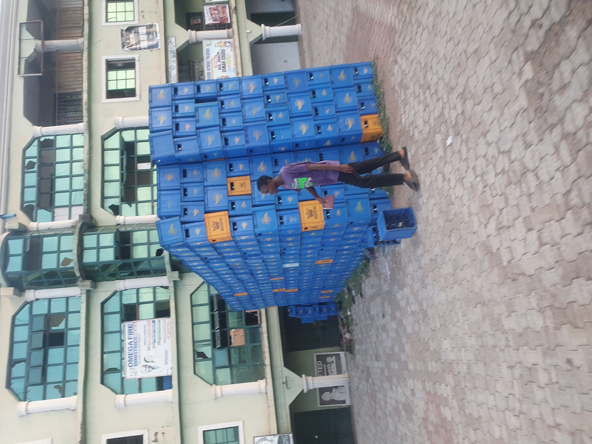 The image, taken in Lokoja, Nigeria, captures a daytime scene featuring a multi-story building and a large stack of plastic crates. A young man walks alongside the towering stack, his attention directed downwards towards an object he holds in his hands, possibly a phone or a book.
The main subjects are the man and an extensive wall-like structure composed of blue plastic crates, interspersed with a few yellow ones, stacked several layers high. The crates appear to be empty, suggesting storage or delivery. One crate in the foreground contains a few indiscernible items. The man is wearing a purple t-shirt and dark trousers.
Behind the crates and the man is a light-colored building with multiple floors. It features various windows, some rectangular and others with an octagonal-like shape, many of which appear to have broken panes. White architectural pillars are visible along the building's facade.
The ground is paved with interlocking brick-like tiles, and a small strip of grass or weeds runs along the base of the crate stack. The lighting suggests it is daytime, possibly early morning or late afternoon, without harsh shadows.
Visible text on banners and posters attached to the building includes "OMEGAFIRE MINISTRIES" with two faces below it, and another banner with "ALH. YAHAYA USMAN ODODO". Another poster is partially visible with "BUILDING REVIVAL PRAYERS". Text or logos on the crates are not clearly legible.