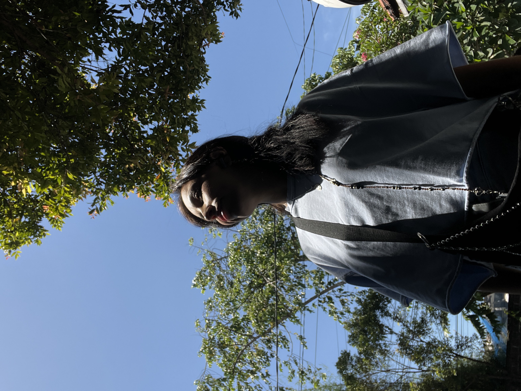 A medium-shot, low-angle photograph shows a woman standing outside on a sunny day in Kuta Selatan, Indonesia. The woman, who is partially visible, has long dark hair and is wearing a light blue top with a black strap. A beaded necklace hangs around her neck.
The background features a clear blue sky partially obscured by lush green foliage from trees. Electrical wires are also visible against the sky. Some buildings can be seen in the distant background. The lighting suggests it is daytime, with the sun positioned high in the sky.