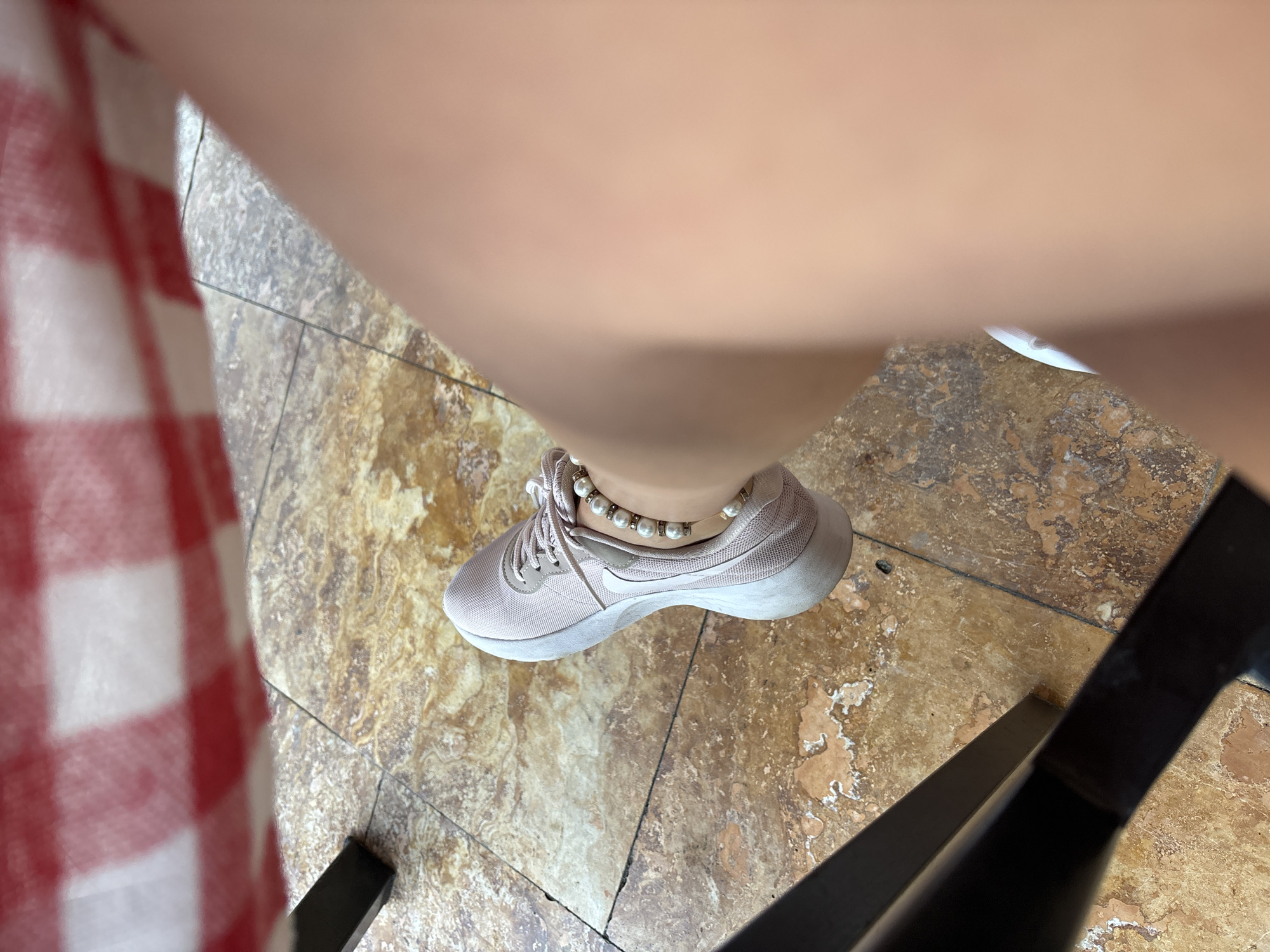 A close-up shot shows a person's leg and foot wearing a light pink sneaker and a pearl anklet. The sneaker is laced and rests on a tiled floor with a marbled pattern. A portion of a chair leg, appearing dark and metallic, is visible in the lower left corner. Above, a red and white checkered tablecloth hangs over the edge of a surface, suggesting a dining setting. The overall impression is a snapshot taken from a seated position, possibly at a cafe or restaurant in Budva, Montenegro, given the typical tile and tablecloth combinations seen in such establishments in the region.