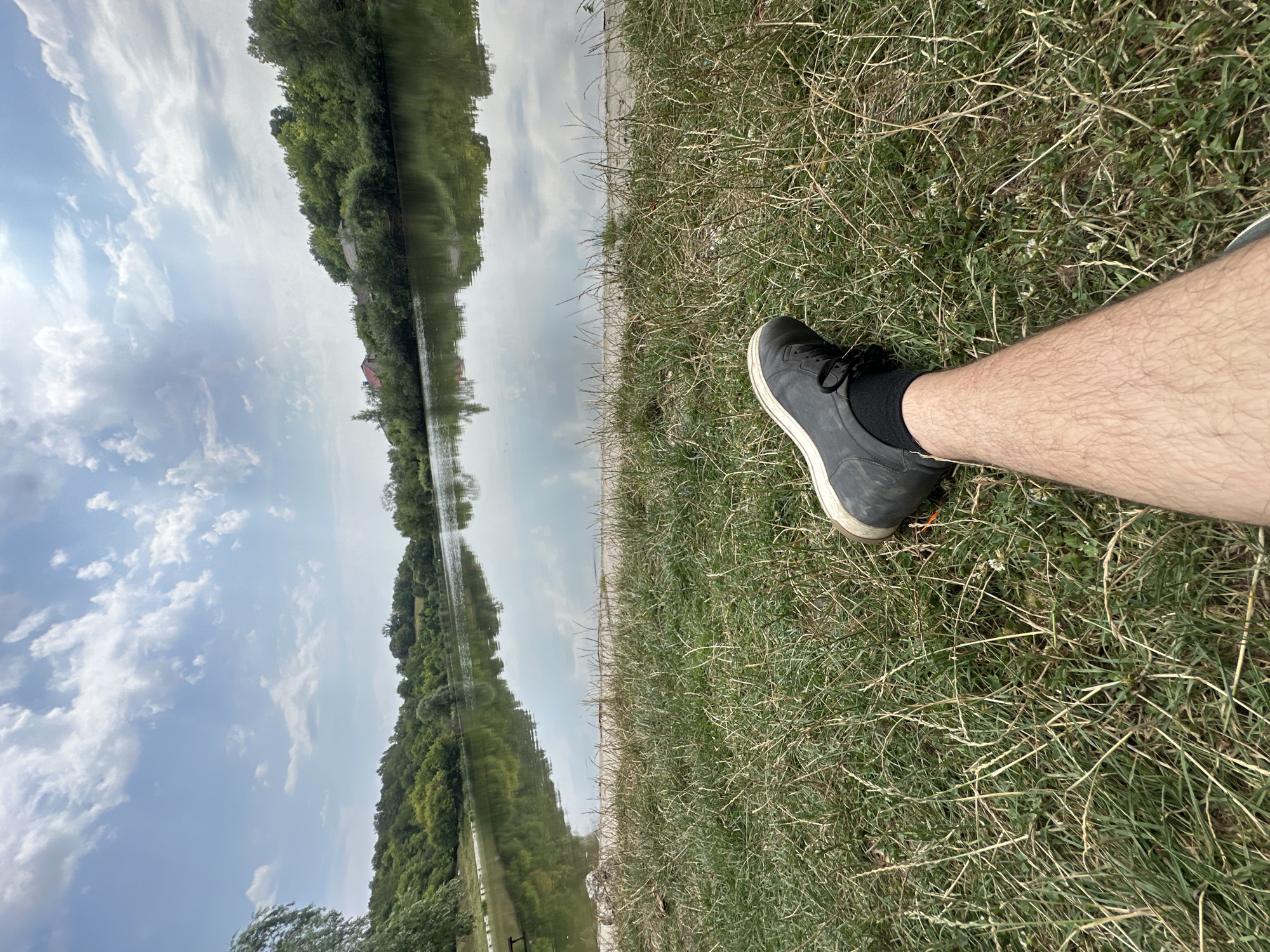 This is a first-person perspective shot of a person's leg and foot resting on a grassy bank next to a calm body of water. The person is wearing dark-colored shoes and socks. The background features a tree-lined shore with some houses visible in the distance, reflecting in the water. The sky is partly cloudy. The scene is set in Makiv, Ukraine.