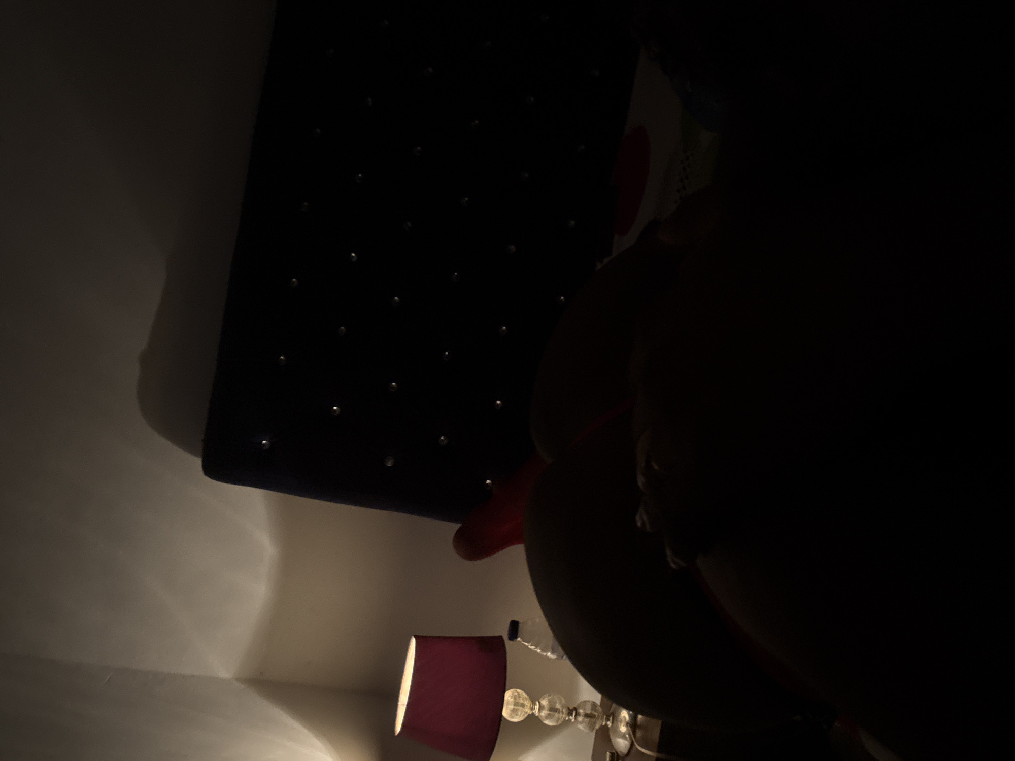 The image depicts a dimly lit room with a dark-colored, studded headboard positioned against a white wall. A small, maroon lampshade is partially visible, illuminating the wall adjacent to it. A clear, ornate lamp base can be discerned beneath the shade. In the foreground, there is a dark, indistinct shape, possibly a person or object, obscuring a significant portion of the view. It is impossible to determine if this image was taken in Abuja, Nigeria, or to provide further detail on the subject.