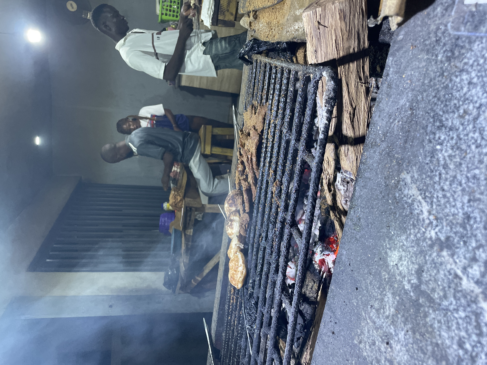 A photograph taken in Akwanga, Nigeria, at night shows a street food scene focused on a large charcoal grill loaded with meat being cooked. The grill is positioned on the right, with charred wood supporting it and a grey concrete surface to its right. In the background, three people are visible: one man in a white shirt holding skewers of meat stands at the top of the frame, while two others are near the grill. One of them is wearing a grey shirt. Smoke rises from the grill.