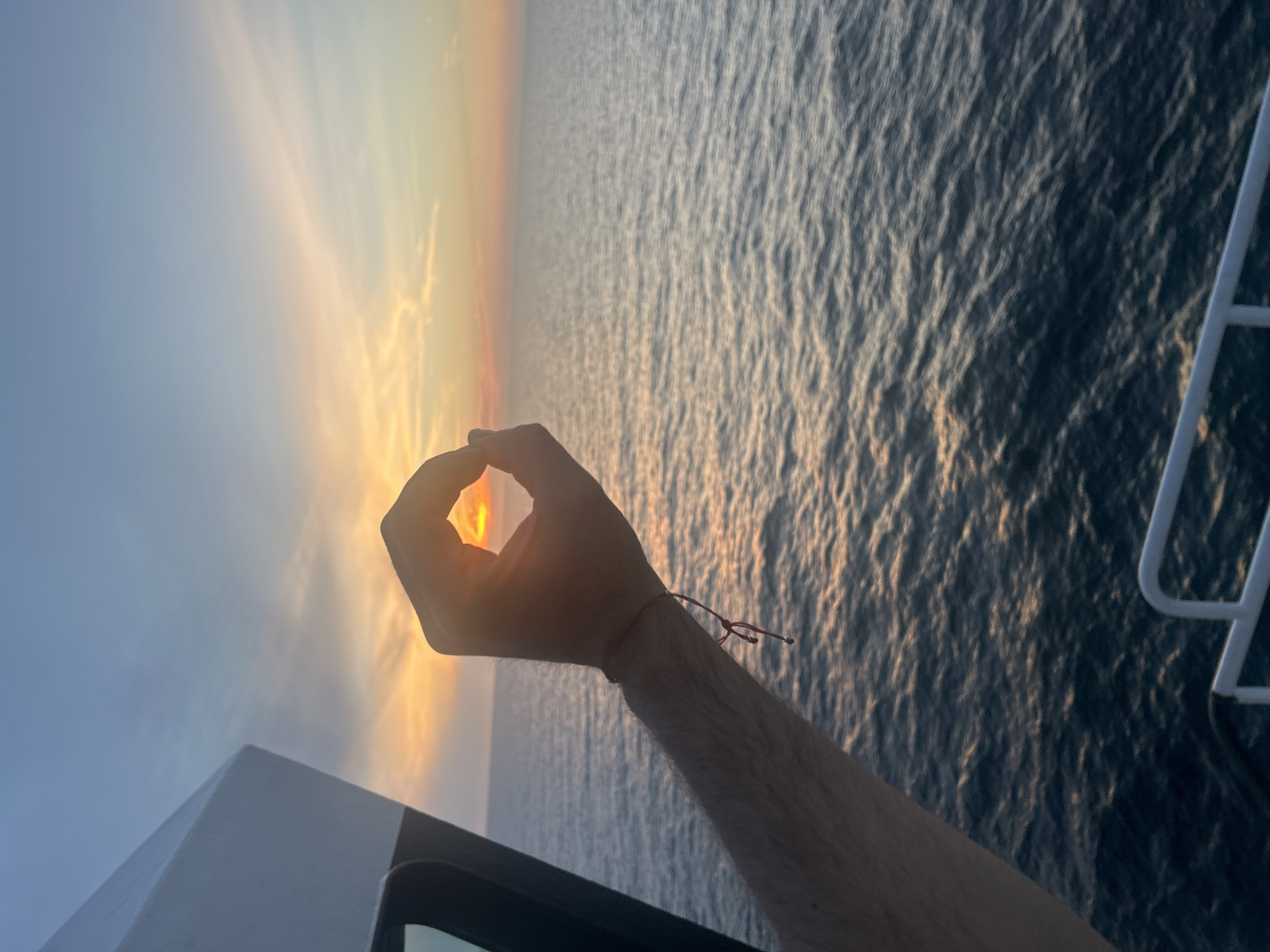 An over-the-shoulder, medium shot captures a person's arm extended towards the left, with their hand forming an "okay" symbol around a setting sun over a body of water. The sky is a gradient of pale blue to orange and yellow, with wispy clouds. The water is dark blue and appears to be choppy, with streaks of sunlight reflecting off the surface. A white railing and a portion of a boat's structure are visible on the left and bottom of the frame, suggesting the vantage point is from a vessel at sea. The person has a thin red string bracelet on their wrist.