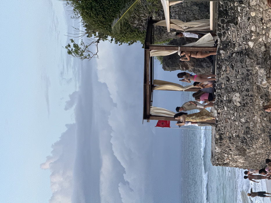 The image portrays a coastal scene in Pecatu, Indonesia, featuring a stone-constructed platform overlooking the ocean. Approximately twelve individuals are gathered on this platform. A woman in a long, light-colored patterned dress stands near a red flag displaying a white circular logo on a pole, actively holding a smartphone as if taking a photograph. Adjacent to her, a man in a beige shirt and shorts is positioned. Further to the right, two women in swimwear, one in a dark bikini and another in a brown bikini, are also using smartphones to photograph the ocean view. Other individuals are seated or standing on the platform, some interacting or observing. Two open wooden pergolas with light-colored drapes are erected on the platform. Below the platform, on a sandy beach, several people are present, with at least two males wading in the shallow ocean water where moderate waves are visible. The background consists of a cloudy sky and a vegetated cliff face to the right.