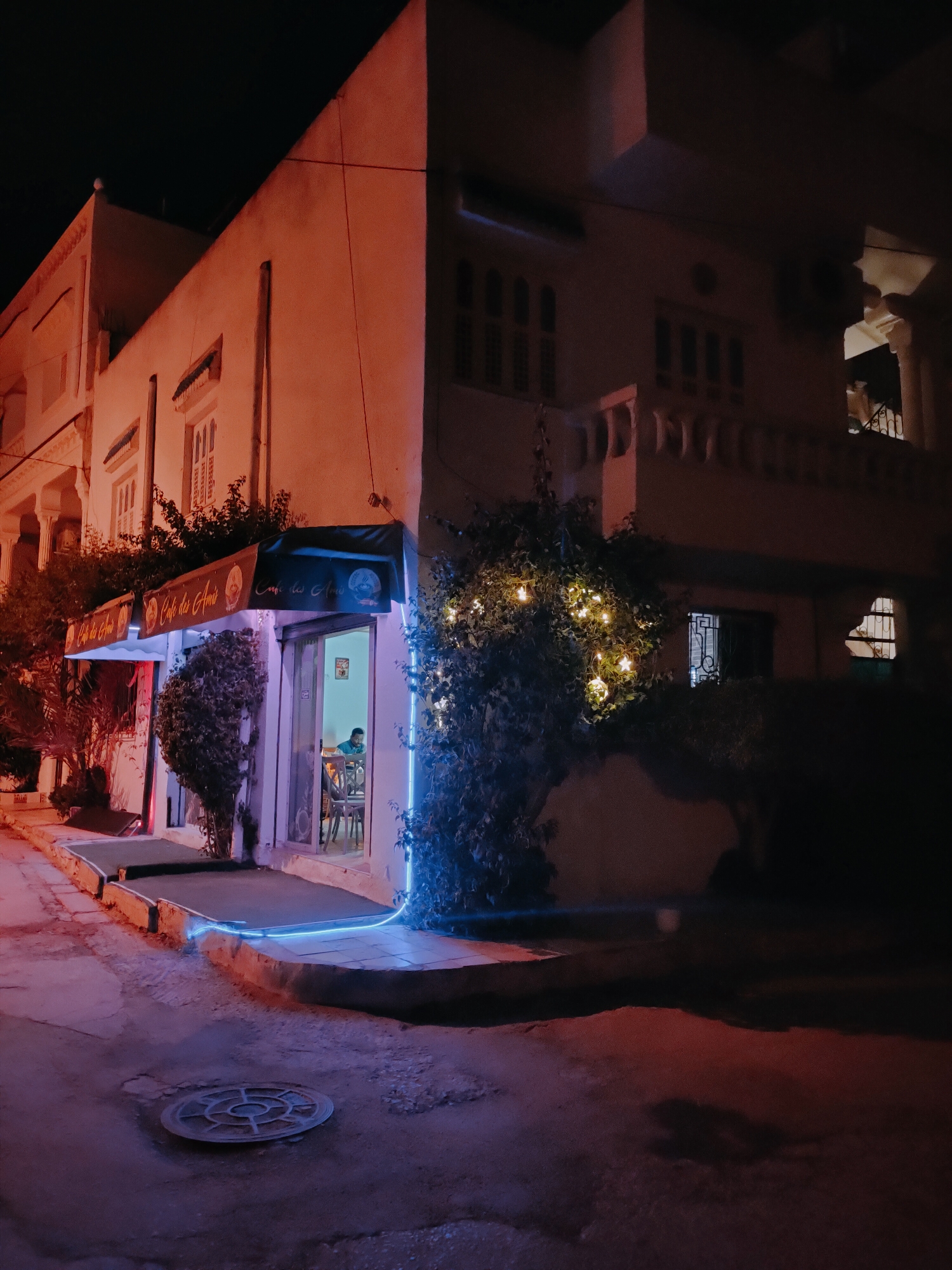 The image depicts a dimly lit street corner in the city of Tunis, Tunisia, at night. A building with a cream-colored facade dominates the scene, illuminated by a mixture of warm orange and cool blue lights. A cafe, identified by the awning displaying "Cafe des Amis," is situated on the ground floor. Through the glass facade of the cafe, a person can be seen sitting at a table, seemingly alone. Lush green foliage surrounds the entrance of the cafe, adorned with a string of warm white fairy lights, creating a festive atmosphere. The street itself is paved and appears wet, reflecting the ambient lights and contributing to the moody ambiance. A circular manhole cover is visible on the road surface. The overall impression is one of a quiet, intimate evening at a local establishment in Tunis.