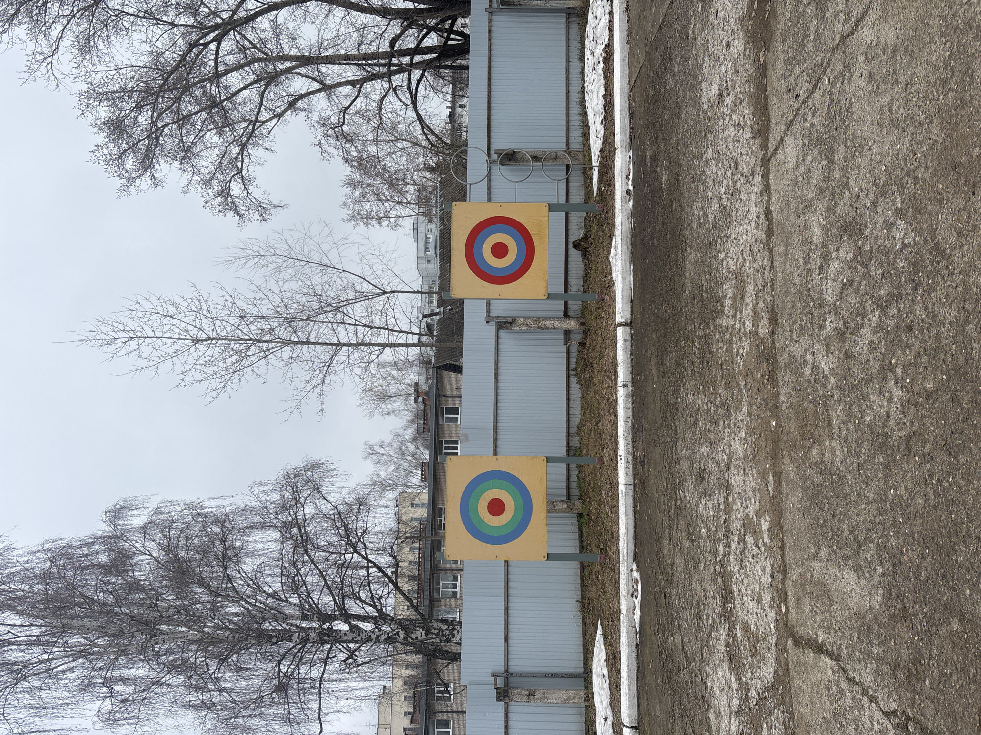 This outdoor image, captured in Chaikovsky, Russia, presents a stark scene on an overcast day, likely in late autumn or early spring. The main subjects are two brightly colored archery targets positioned against a backdrop of bare trees and a gray fence.
The target on the left features a red bullseye surrounded by concentric rings of green, blue, and an outermost yellow. The target on the right has a red bullseye with blue and yellow concentric rings. Both targets are square and mounted on simple metal stands.
Behind the targets runs a tall, corrugated gray metal fence, beyond which several multi-story buildings with numerous windows are visible through the leafless branches of trees. The ground in the foreground and to the right is a dark, rough concrete or asphalt path, bordered by a white curb. Patches of snow and ice are visible along the curb and at the base of the fence, indicating cold weather. The sky is a uniform, dull gray, with no direct sunlight.
Numerous bare deciduous trees fill the upper and left sections of the frame, their intricate branch patterns starkly outlined against the pale sky. The overall scene suggests a functional outdoor area, possibly a schoolyard or community space, used for target practice. There are no people visible, and no discernible text is present in the image.