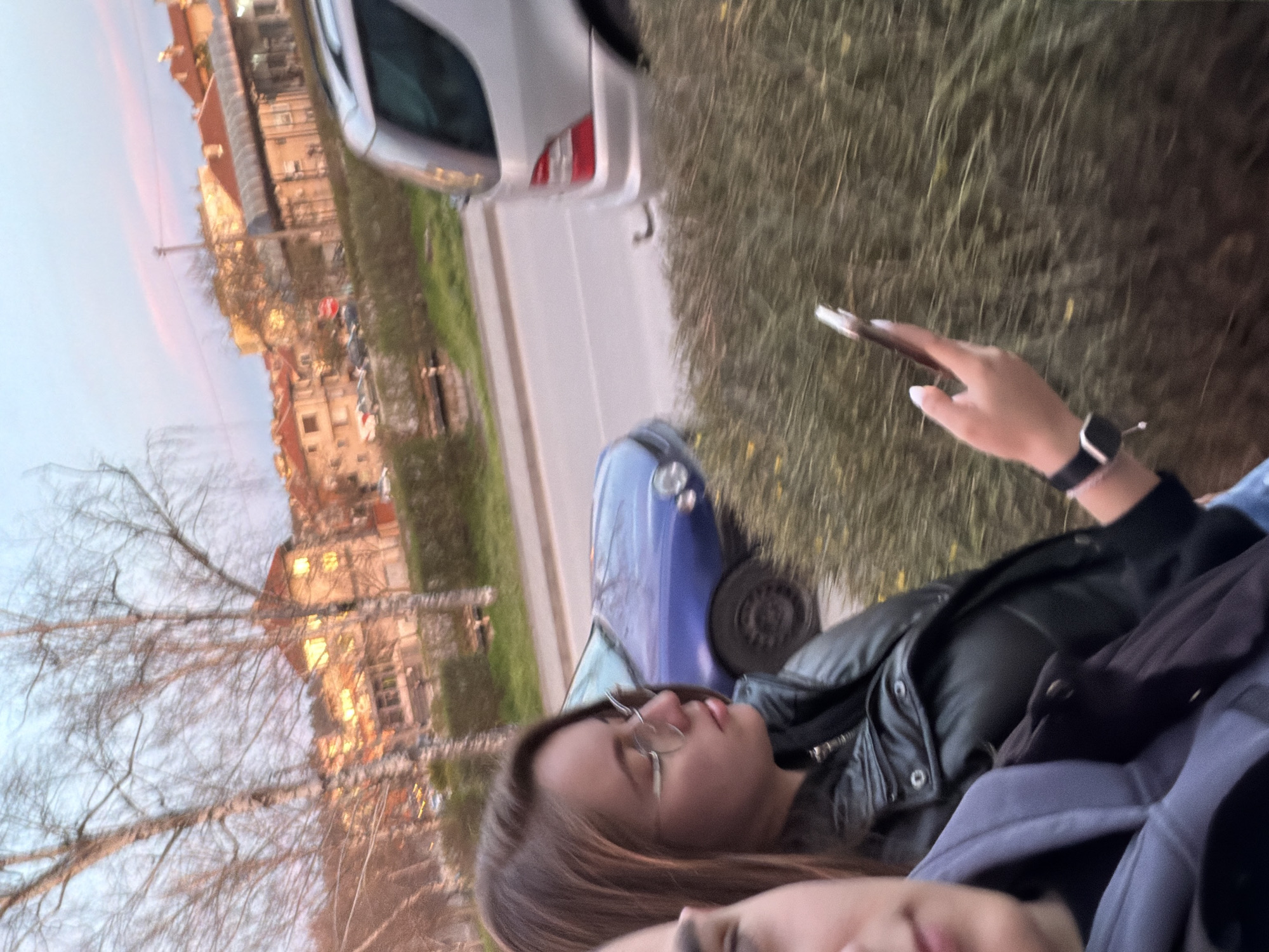 Two individuals are in the foreground. One is holding a smartphone. Behind them is a street with a silver-colored car and a blue car. Buildings are in the background, consistent with an urban setting. The location is Panagyurishte, Bulgaria.