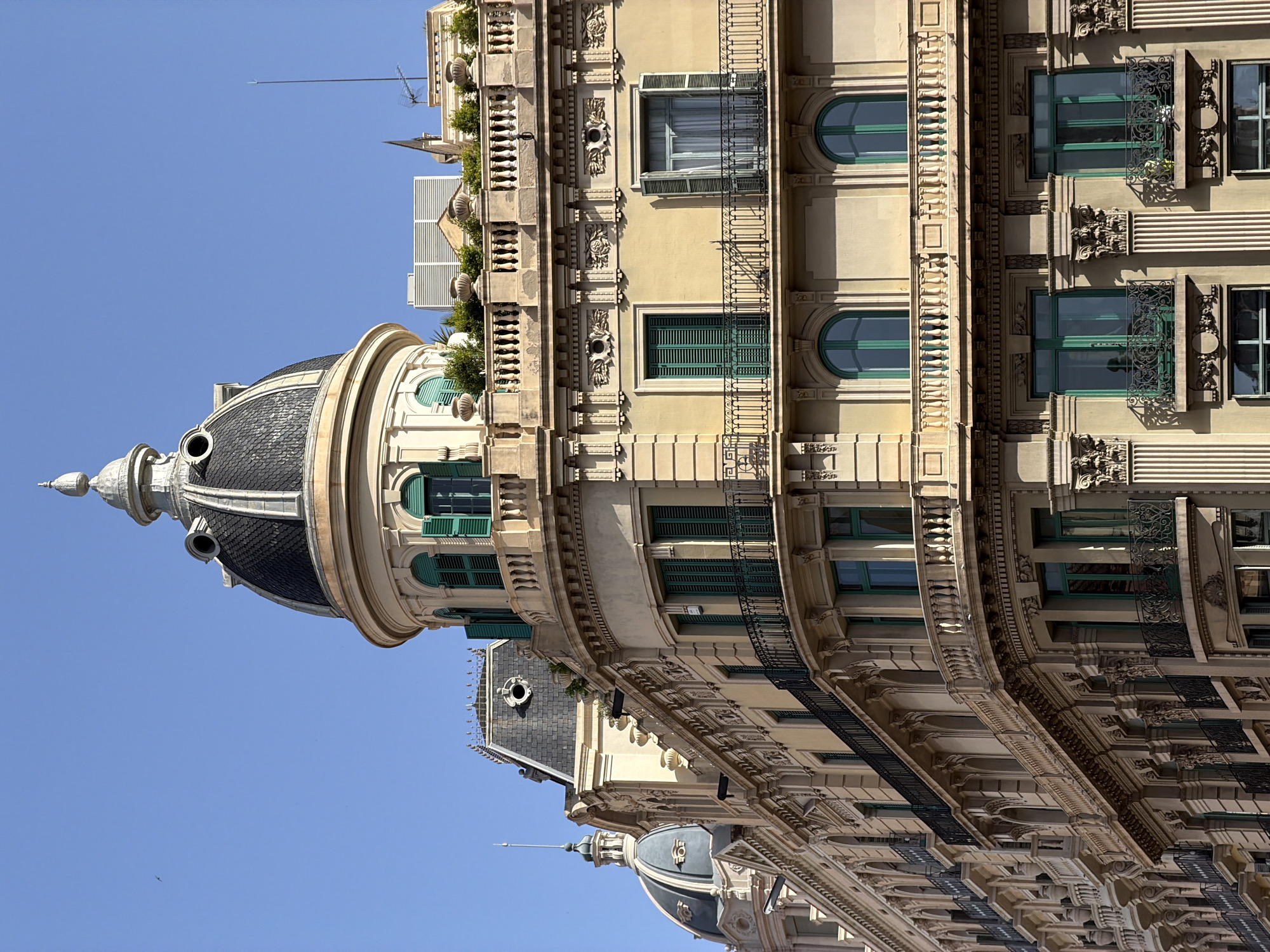 This is a close-up, eye-level photograph of the upper floors of ornate buildings in Barcelona, Spain, under a clear blue sky. The architecture is classical with Baroque influences. The prominent building on the right features a curved facade with a large dome topped by a finial, detailed decorative moldings, and a balustrade-lined terrace with potted plants. It has multiple levels of windows with green shutters and intricate wrought-iron balconies. Another domed building, similar in style, is visible to the left, partially obscured by the main building. The structures appear to be residential or commercial properties. The lighting suggests it is daytime, with sunlight casting shadows on the building's facade, highlighting its architectural details.