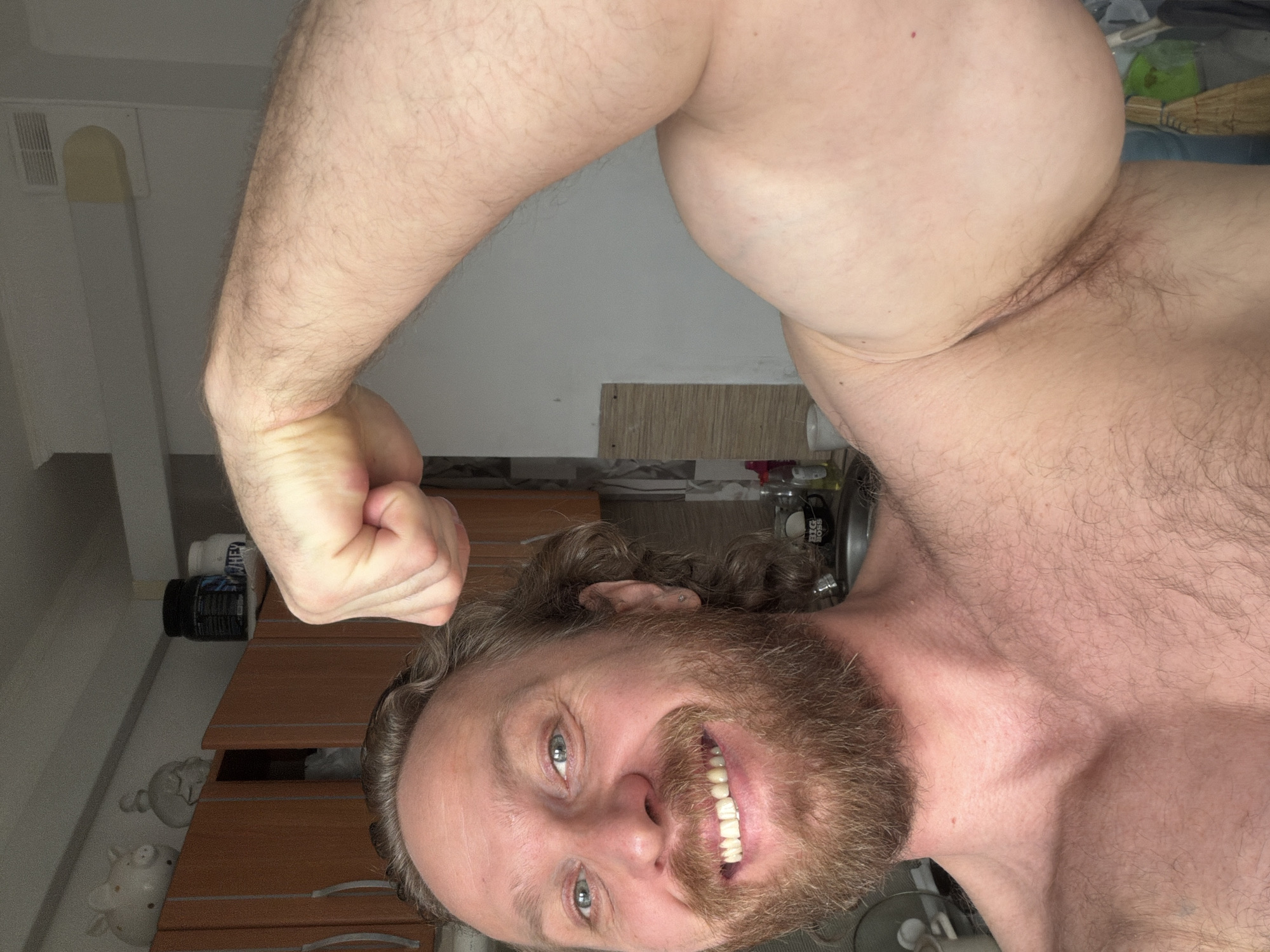 A shirtless man with a beard is smiling at the camera and flexing his right arm. He appears to be in a domestic setting, possibly a kitchen or bathroom, as suggested by the presence of cabinets, a supplement container, and other household items in the background. There is no indication of the location being Saint Petersburg, Russia.