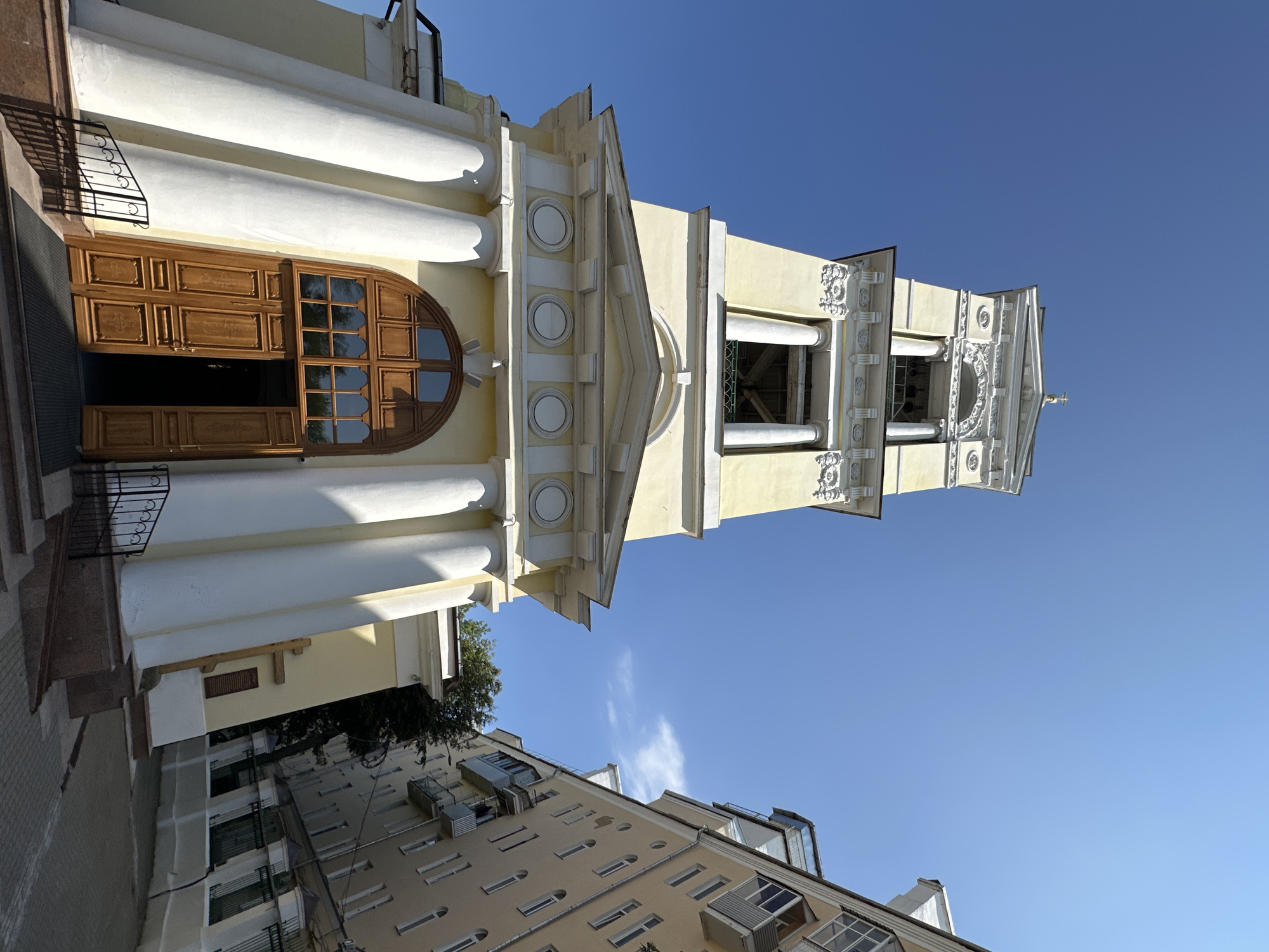 The image depicts the bell tower of the Orthodox Church of the Resurrection of Christ in Podolsk, Russia, viewed from a low angle against a clear blue sky. The bell tower is painted a pale yellow and features white classical architectural elements, including columns, cornices, and decorative molding. A large, arched wooden double door is visible at the base of the tower, with one door ajar. Adjacent to the church, on the right side of the frame, is a multi-story residential building with balconies and windows, also against the bright blue sky. A paved walkway or street is visible leading towards the church entrance and between the buildings. The overall scene is one of urban architecture under a sunny sky.
