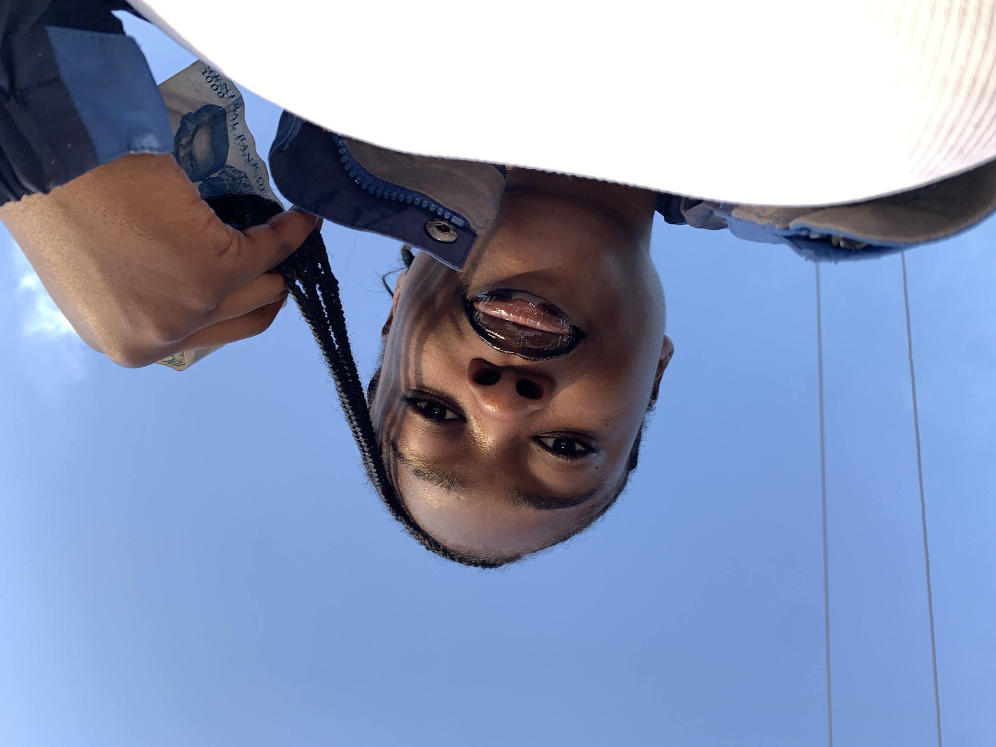 In Rafin Par, Nigeria, a low-angle outdoor shot captures a person's face from a worm's-eye view against a clear blue sky. The subject, with braided hair pulled back and adorned with dark lipstick, looks directly at the camera. A hand is visible, holding a banknote with text that appears to be "1000" and "BANK OF". The person is wearing a blue and gray jacket. The image is tilted, with two thin white wires running horizontally across the lower part of the frame.