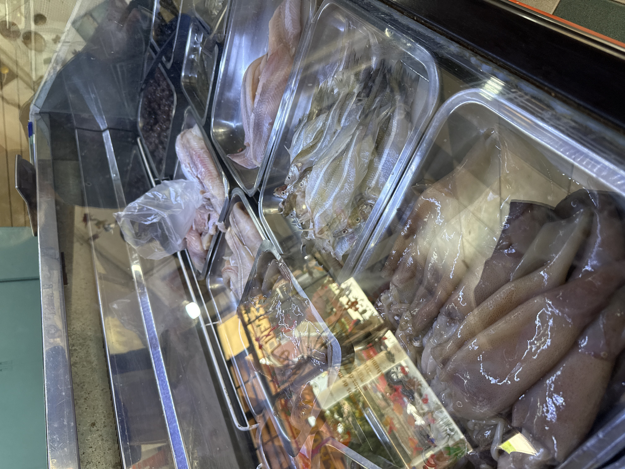 A refrigerated display case containing various raw seafood items in plastic containers is shown. From left to right, the containers hold small, dark items (possibly olives or anchovies), fish fillets, small whole fish, and squid. The display case is located in Villa General Belgrano, Argentina. The environment is a marketplace or food retail establishment.