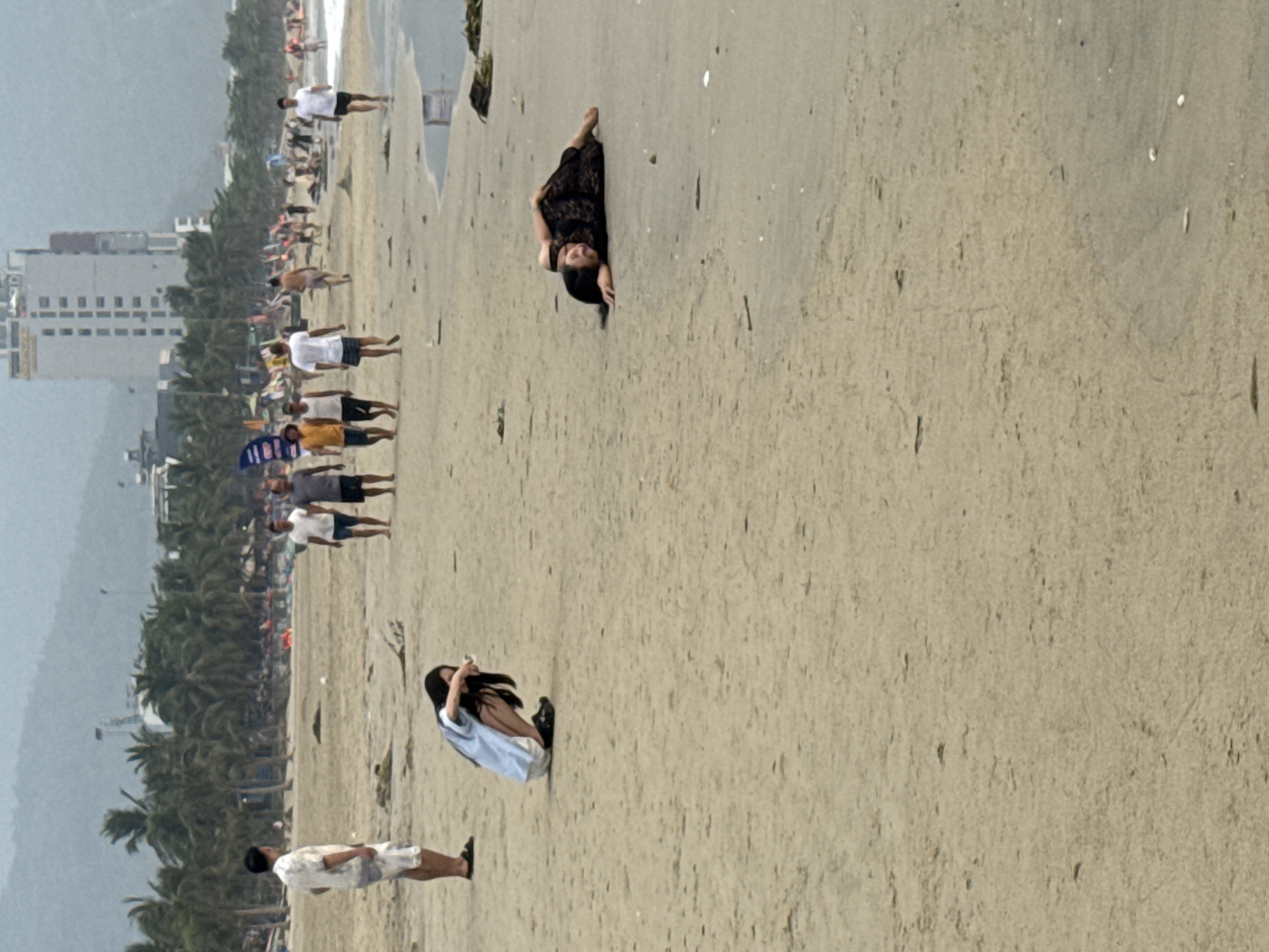 The image shows a beach scene, likely in Đà Nẵng, Vietnam, given the presence of high-rise buildings, palm trees, and a crowded beach. Several people are scattered along the beach; some are walking, and others are sitting or lying down.
Specific individuals are identifiable: one person is lying prone on the sand in a black dress, and another person is seated and facing away from the camera wearing a light blue top. A man stands toward the bottom of the image, wearing a short-sleeved, patterned shirt and shorts. He faces away from the viewer, and the beach appears to stretch out behind him.
The environment includes a wide sandy beach, a body of water (ocean), and a cityscape with buildings and palm trees extending along the coastline. The overall lighting suggests a sunny day.