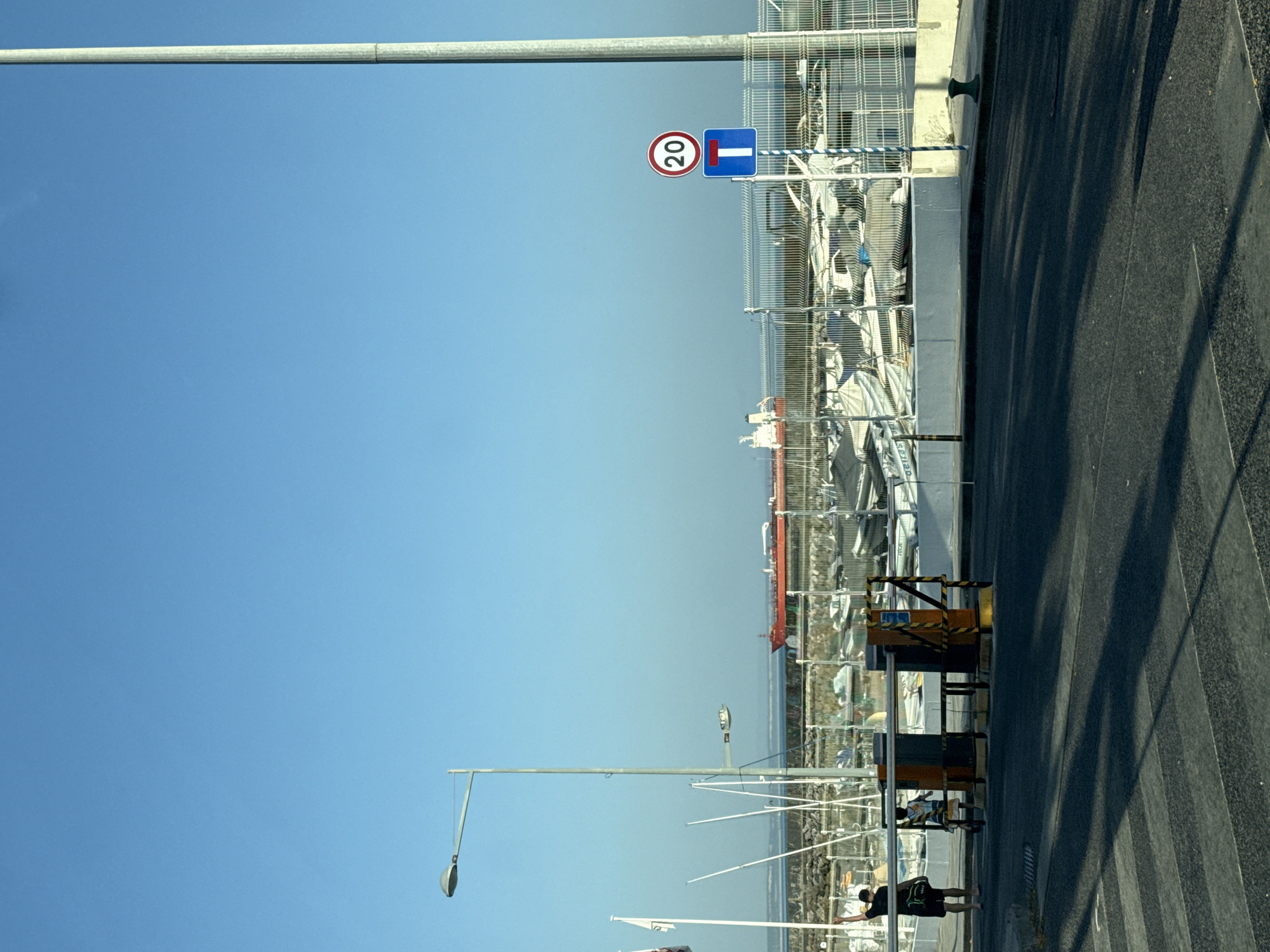 The image captures a street scene in Oeiras, Portugal, on a clear day with a bright blue sky. In the foreground, a crosswalk is visible on dark asphalt. A striped barrier gate is positioned on the right side of the road. Two individuals are present near the gate: one adult wearing dark clothing and a bag over their shoulder, and a child in a patterned shirt. Behind the gate, a large red ship is anchored in the water, with numerous smaller boats moored in a marina. A tall lamppost stands to the right of the frame, and a traffic sign displaying a speed limit of 20 is mounted on a pole. A metal fence separates the road area from the marina. The overall environment suggests a coastal town with maritime activity.