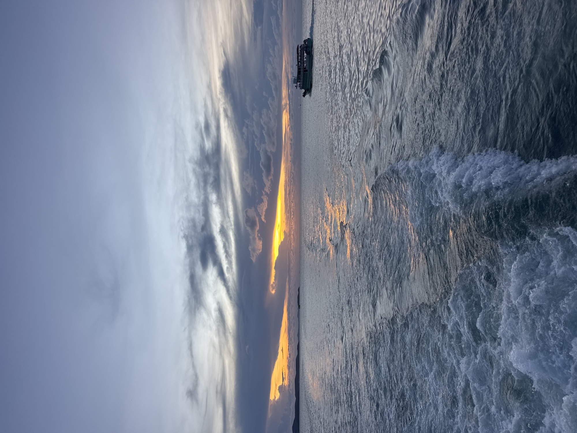The image depicts a body of water, likely the sea, with a sunset reflecting on its surface. A boat is visible on the water. The sky is filled with clouds, and the sun is setting on the horizon. The location is possibly Pattaya City, Thailand.