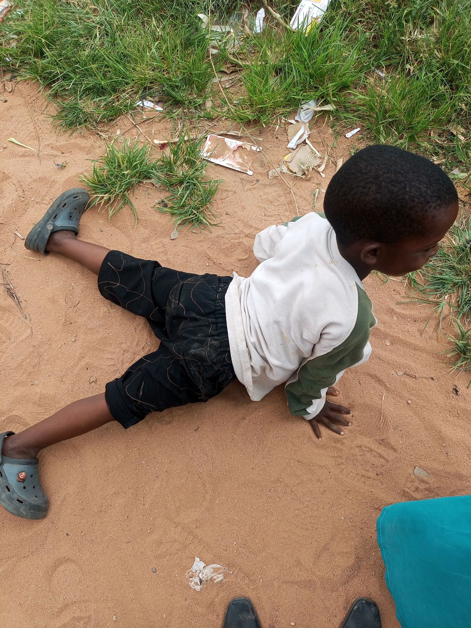 A young boy with dark skin and short, dark hair lies on sandy ground, his body stretched out with his left leg extended. He wears black shorts with a subtle pattern and black Croc-style shoes. His upper body is clad in a white long-sleeved shirt with green accents on the sleeves and collar. His right hand is extended, palm down, on the sand. The environment is characterized by dry, sandy earth interspersed with patches of dry grass and scattered debris. The scene appears to be outdoors, possibly in a semi-urban or rural setting within Tempure, Nigeria, given the arid ground and vegetation. The lighting suggests daylight.