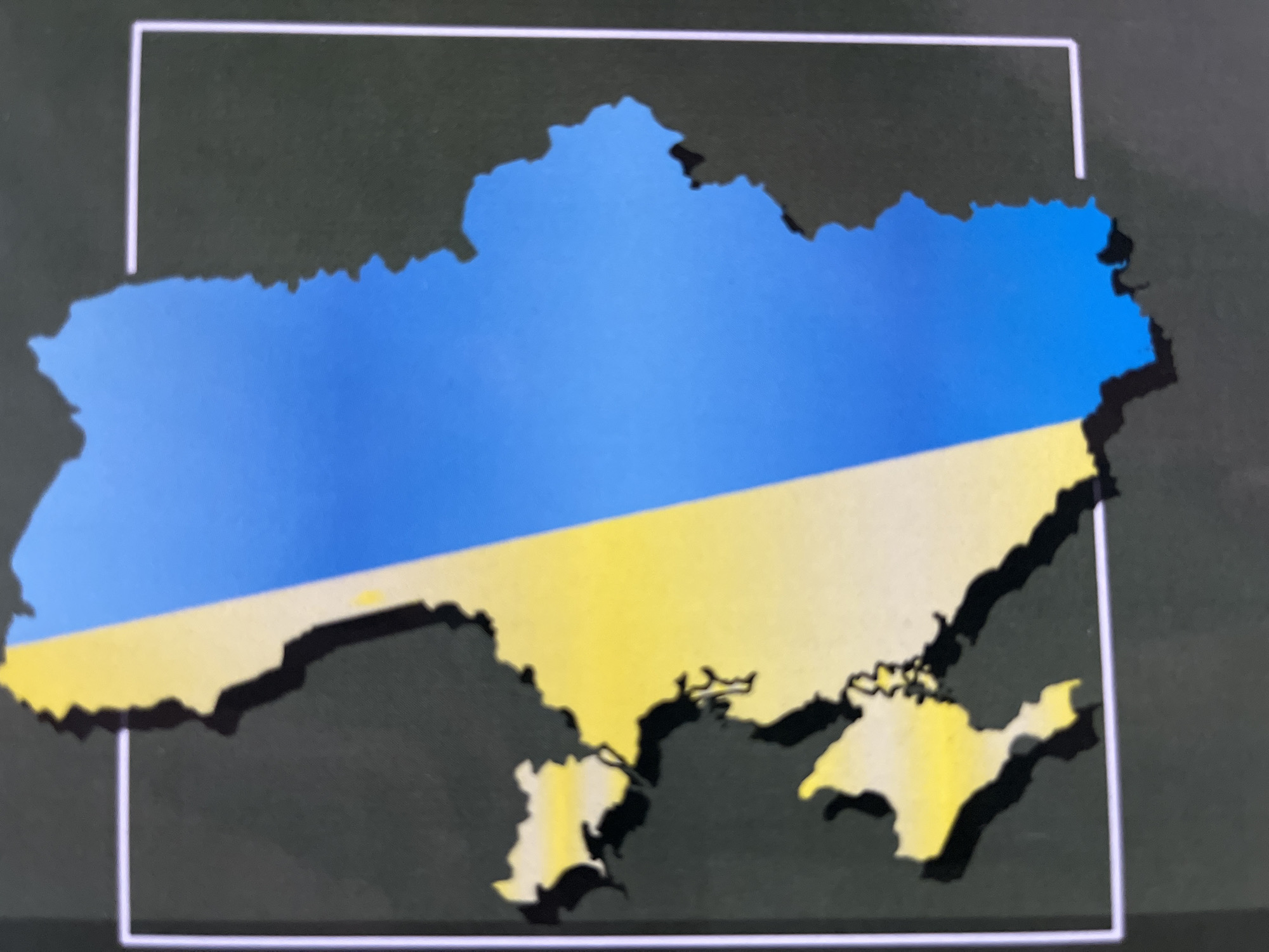 The image displays a stylized map of Ukraine, filled with the colors of its national flag. The upper portion of the map is a vibrant blue, transitioning diagonally to a bright yellow in the lower section, mimicking the blue sky over a field of wheat. The geographical contours of Ukraine, including the Crimean Peninsula which appears in the yellow portion, are clearly visible. A subtle, dark shadow effect underneath the map gives it a slightly raised appearance. The map is set against a dark gray background. A partial white rectangular outline, appearing as two L-shaped corners, frames the map. No activity is depicted, and there is no visible text. The image appears to be a photograph of a digital display or a printed graphic.