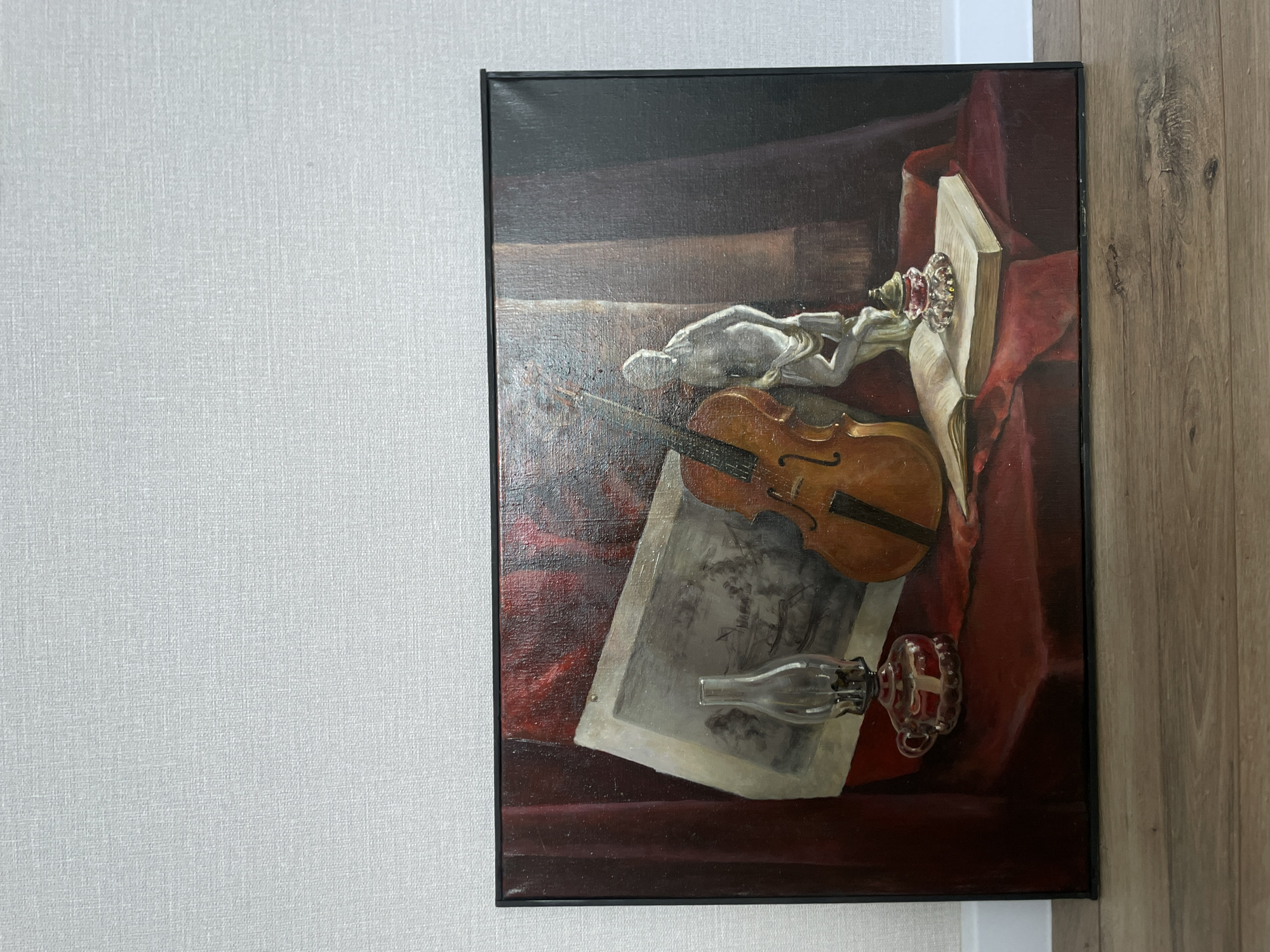 The image shows a still-life painting hanging on a wall. The painting depicts a violin, a statue of a reclining figure, a book, a glass object, and a piece of paper, all arranged on a dark red surface. The painting is framed in black. The wall behind the painting is textured and light gray on the left side, transitioning to a white corner and then to a wooden panel on the right side. No specific information about Подшивалово, Russia, is discernible from the image.