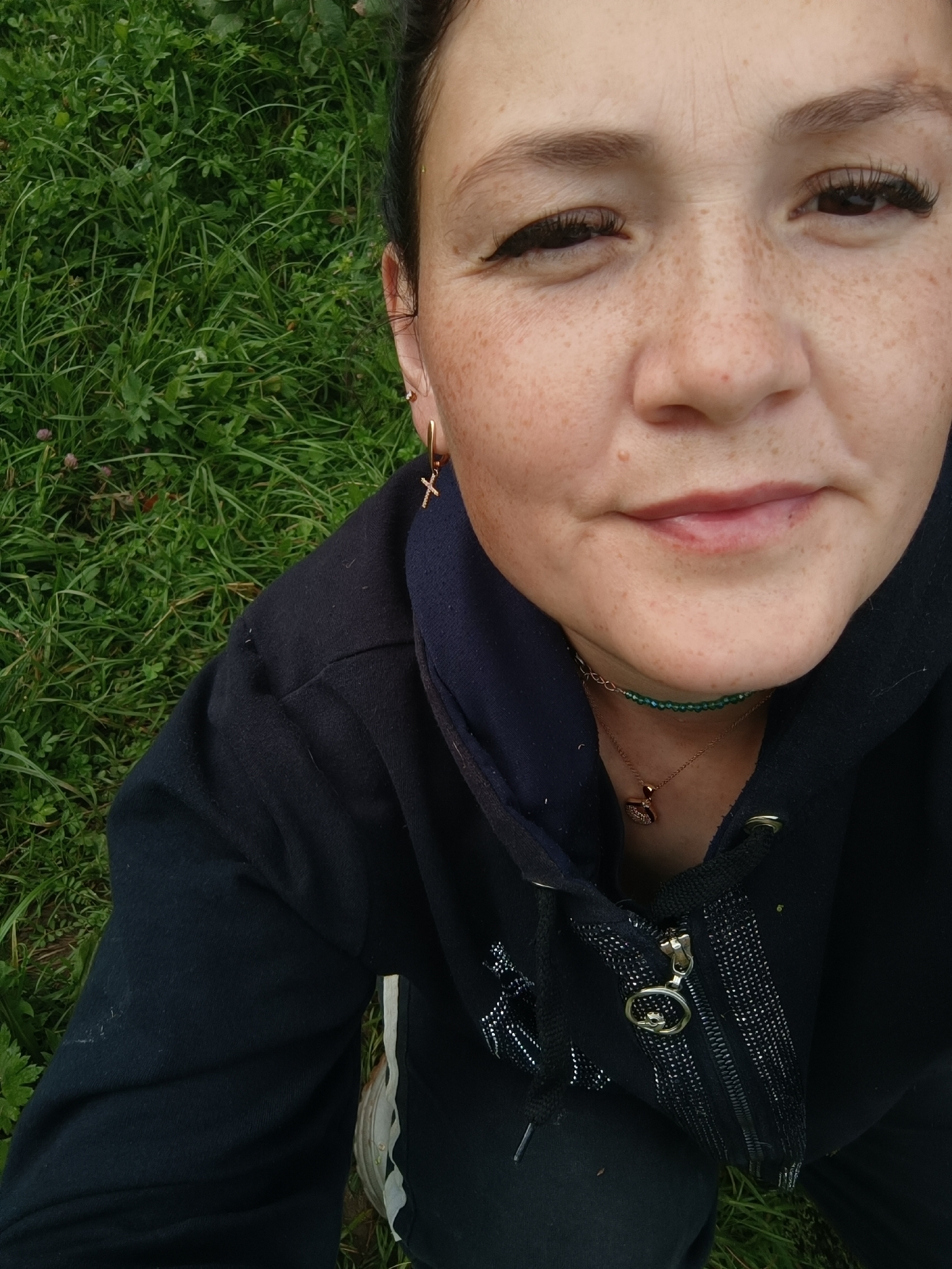 A woman is photographed from a high angle, looking upwards towards the camera. She has dark hair pulled back, and her face is adorned with freckles. Her eyes are wide, and she is wearing noticeable false eyelashes. She has a subtle smile on her lips, and a mole is visible on her upper lip. Around her neck, she wears two necklaces: one a thin strand of green beads, and the other a gold chain with a heart-shaped pendant. She is also wearing a gold cross earring in her visible ear. She is dressed in a dark blue hooded jacket, zipped up. The background is filled with green grass, suggesting an outdoor setting. The image does not contain any information that would allow for the determination of the city of Karsha, Russia.