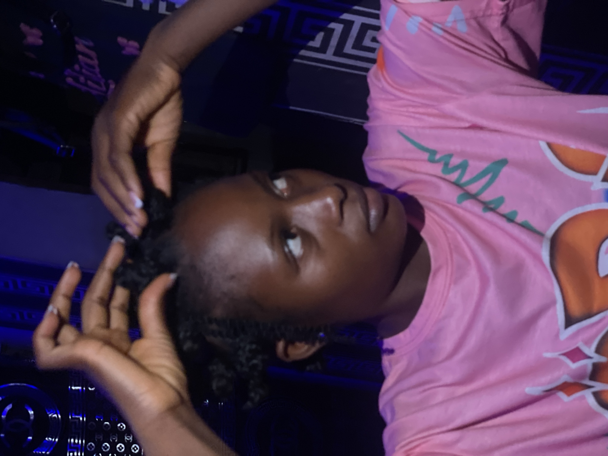 A woman with dark skin and short, tightly curled hair is looking upwards and to the side. She is wearing a pink shirt with a large, cartoon-like design featuring orange and white. Her hands are raised near her head, as though she is styling or touching her hair. The background is dark and indistinct, with what appears to be a patterned surface behind her head and shoulders. The image does not provide enough information to confirm the location in Lokoja, Nigeria.