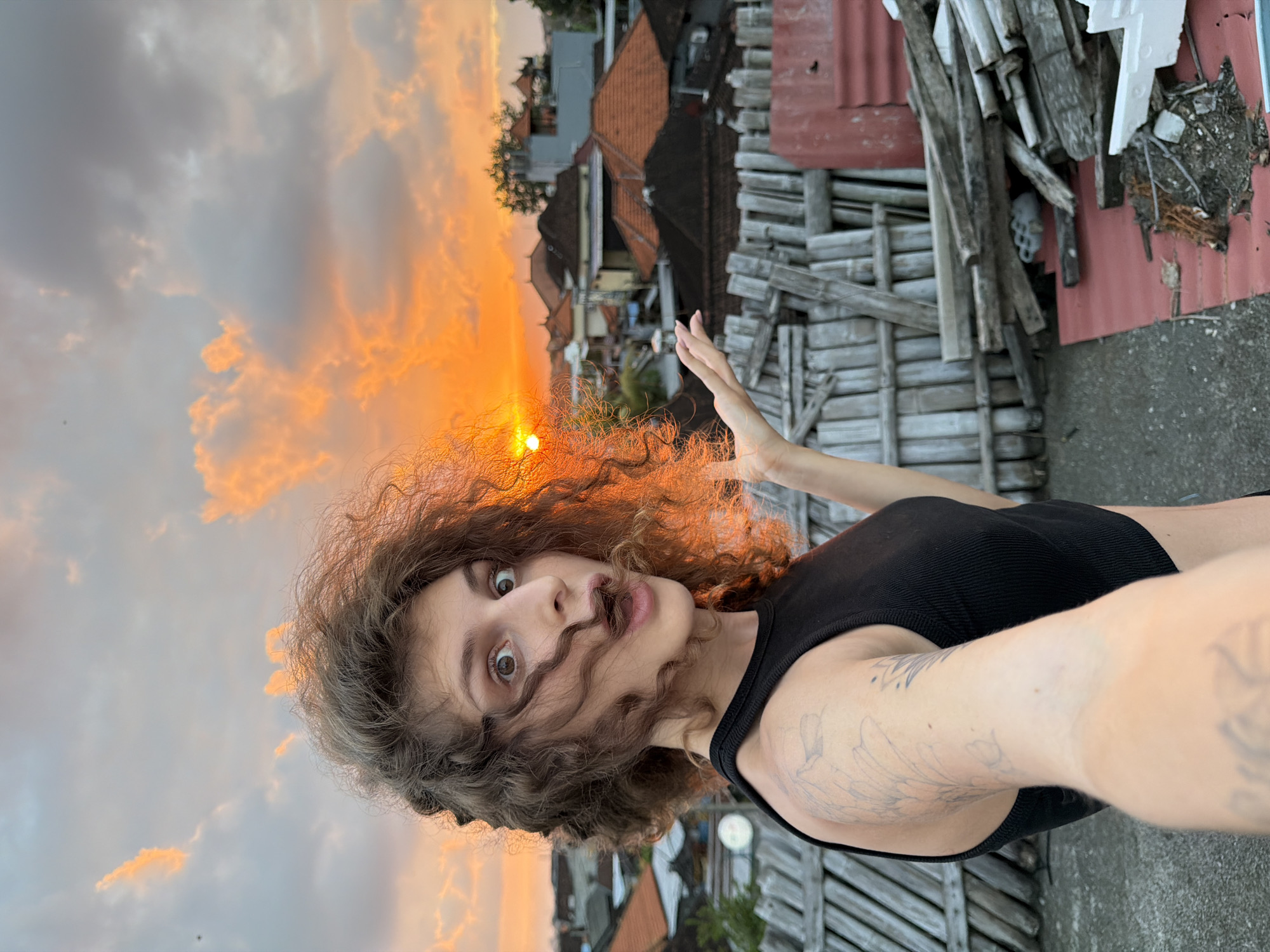 A young Caucasian female with curly brown hair is captured in a selfie taken outdoors at sunset in North Kuta, Indonesia. She is wearing a black sleeveless top and has tattoos on her left arm. Her facial expression is one of surprise or excitement, with her mouth open and eyes wide. Her hair is windswept, with some strands obscuring her face.
The background reveals a cityscape with numerous rooftops and buildings. The sky is filled with dramatic clouds illuminated by the setting sun, casting a warm orange glow. In the foreground, a pile of weathered wooden planks and debris is visible, suggesting a rustic or construction area. The overall lighting indicates that the photo was taken during the golden hour.