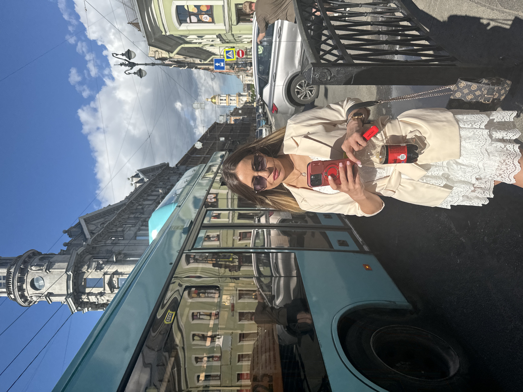 This media file shows a young woman taking a selfie outdoors in what appears to be an urban setting. The image is oriented vertically with the bottom of the frame on the right. The woman is wearing a beige jacket over a white lace dress and sunglasses, with a necklace and bracelet. She is holding a red smartphone in her right hand, and her left hand is holding a small red object, possibly a lipstick or a cosmetic item, and a glass bottle with a label that says "Lost Man". Next to her is a patterned purse with a chain strap.
Behind her, a turquoise bus is prominently featured, its windows reflecting the surrounding buildings and sky. In the background, there are ornate historical buildings with domed structures and intricate architectural details, characteristic of Saint Petersburg, Russia. A silver car is parked on the street, and a man in a brown shirt is visible near it. A black metal fence is in the foreground, partially obscuring the view. Streetlights and overhead wires are also visible.
The sky is blue with scattered white clouds, suggesting it is daytime, likely morning or afternoon, with fair weather. The overall mood seems casual and relaxed, possibly a tourist exploring the city. No explicit text is clearly legible on the buildings or signs in the image, other than the "Lost Man" label on the bottle.