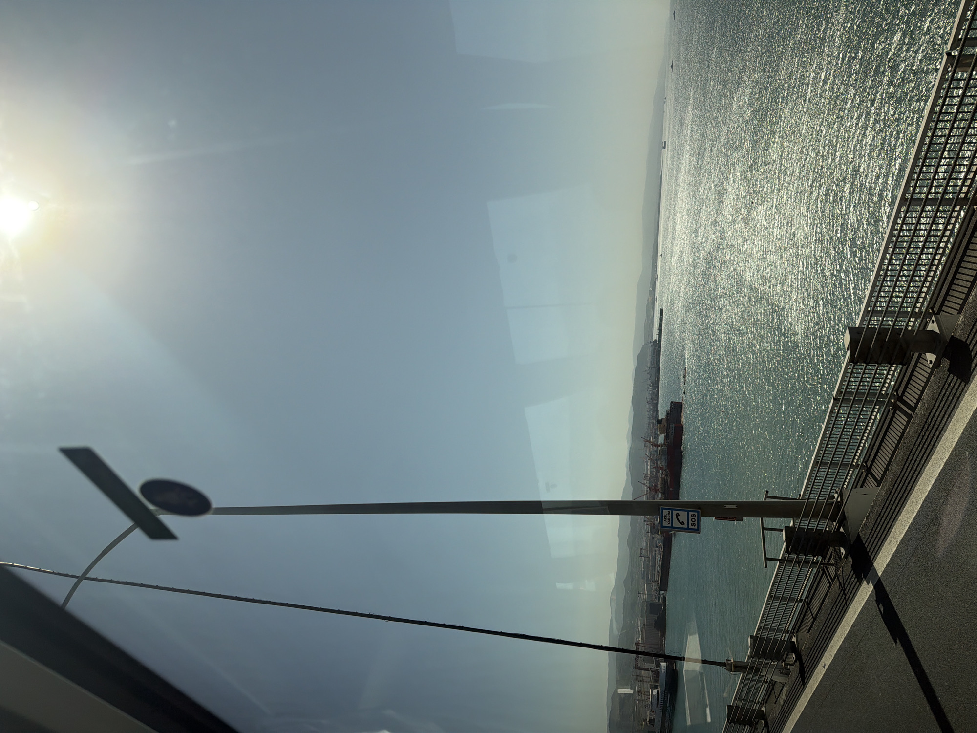 The image displays a sun-drenched, expansive view over a large body of water, likely a bay or sea, from an elevated perspective, possibly a bridge or coastal highway. The bright sun, located in the upper left, casts a strong glare and lens flare, illuminating a clear, light blue sky that dominates the upper half of the frame.
To the right, a road with a robust metal railing runs along the edge of the scene, extending into the distance. The water below glitters with countless reflections of sunlight, indicating calm conditions. Along the distant horizon, rolling hills or mountains are visible, while numerous ships, including large cargo vessels and smaller boats, dot the water. A port or industrial area with cranes and other structures can be discerned on the far shore, suggesting significant maritime activity.
A tall, dark utility pole stands prominently in the foreground, positioned slightly left of center. Attached to this pole is a blue rectangular sign that displays a white phone icon and the text "ACİL YARDIM" above "SOS." This visible text is Turkish for "Emergency Help." Reflections, possibly from a vehicle's window, are subtly visible across the image, particularly in the sky and water areas.
The scene depicts a clear, bright day, likely late morning or early afternoon, given the sun's position. The presence of a bridge-like structure, a busy bay with industrial elements, and the Turkish text on the sign strongly align with the provided context of Dilovası, Turkey.